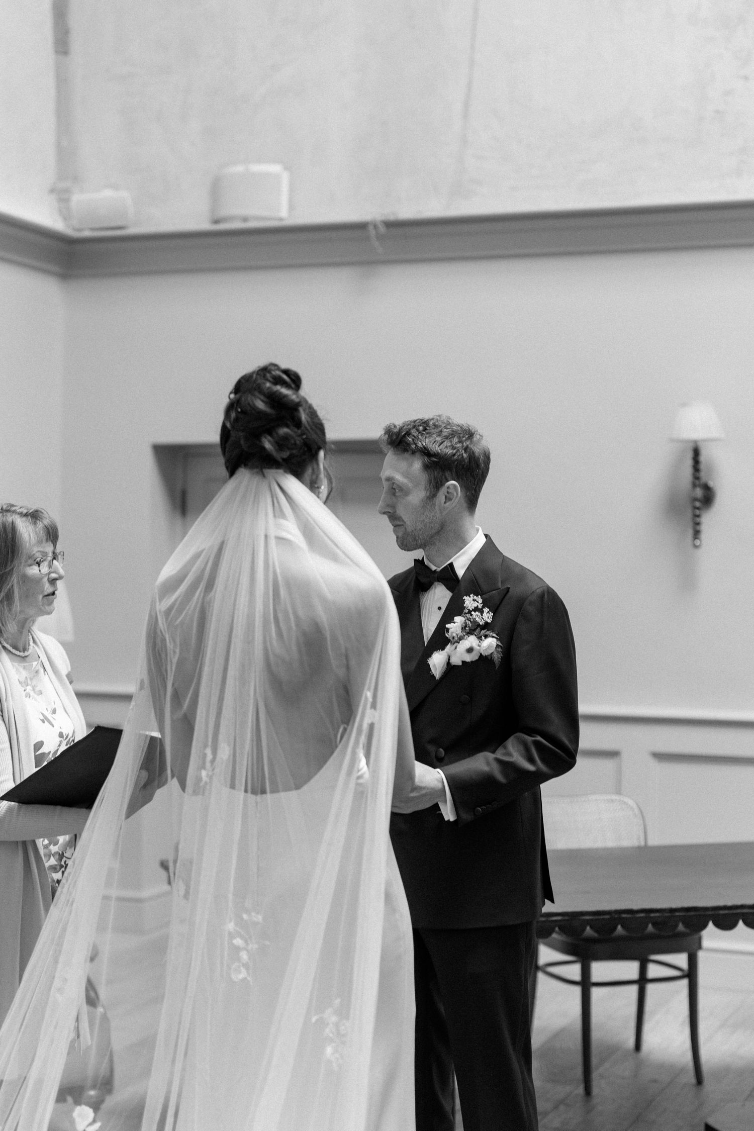 Groom gazing lovingly at his bride during an intimate indoor wedding ceremony with an officiant nearby.