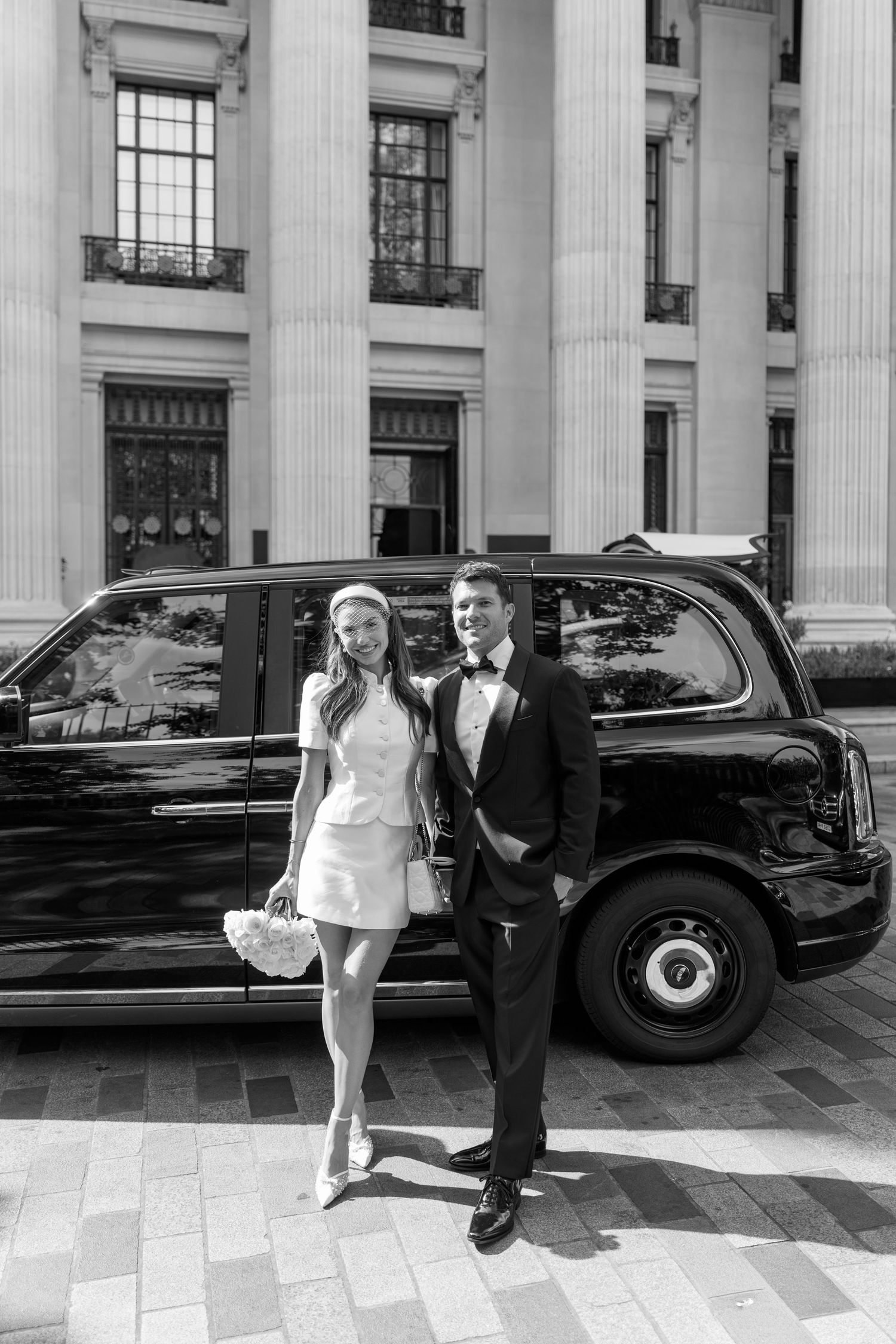 Newlyweds in wedding attire posing beside a classic black London taxi cab outside grand building.