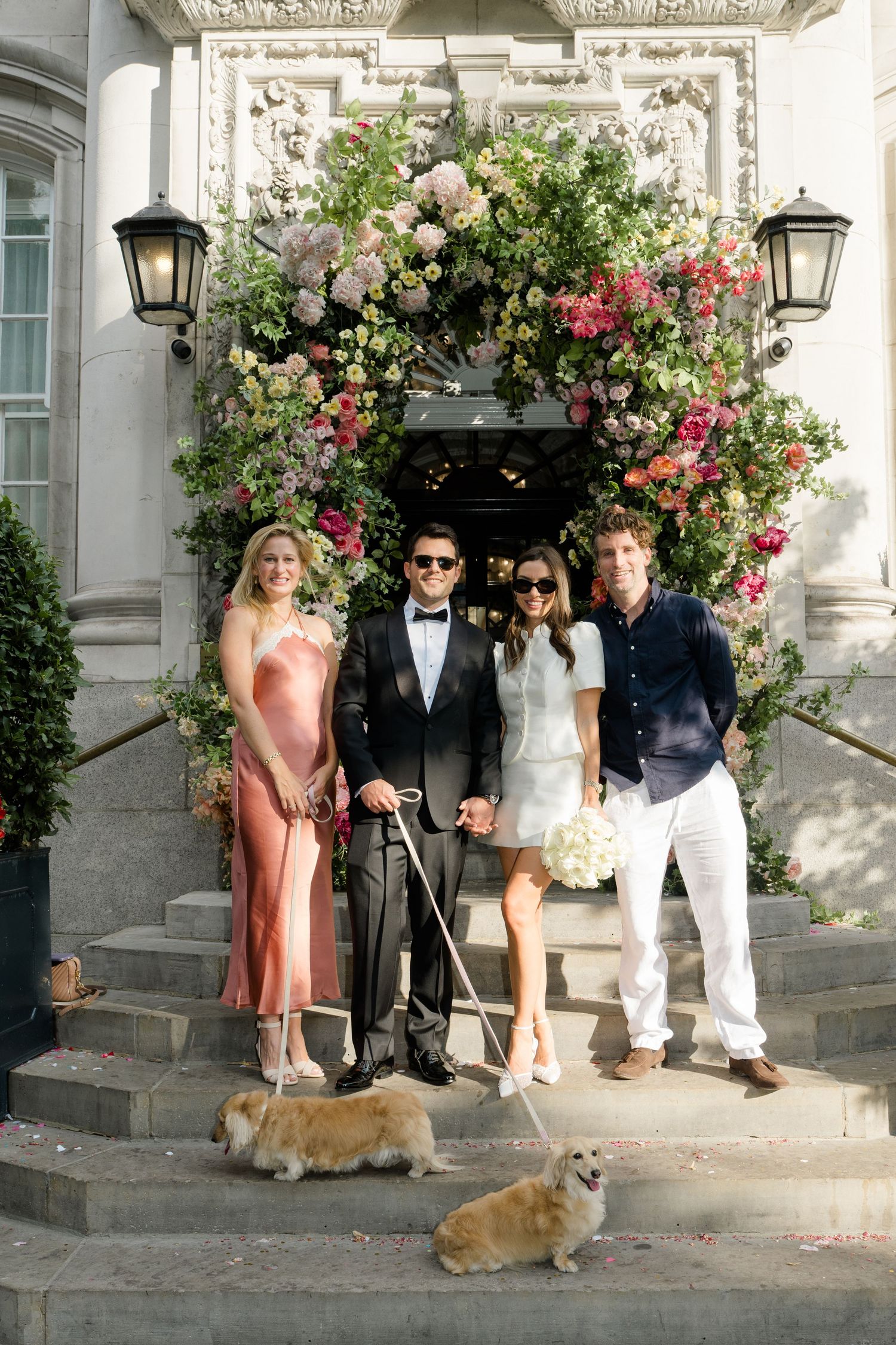 Wedding party of four poses with two corgis under a floral arch.