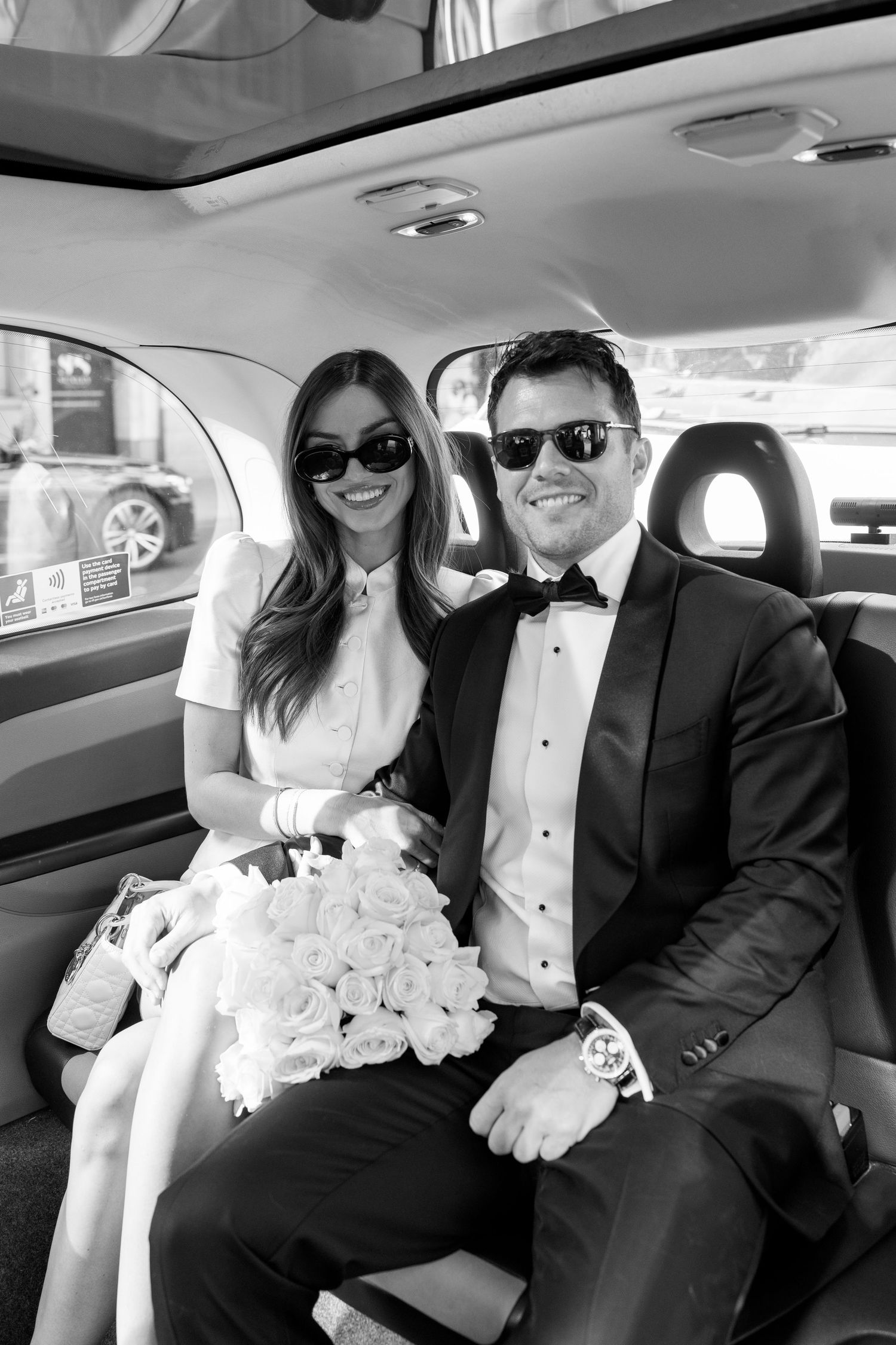 Black and white photo of newlyweds in sunglasses sitting in backseat of car with white rose bouquet.