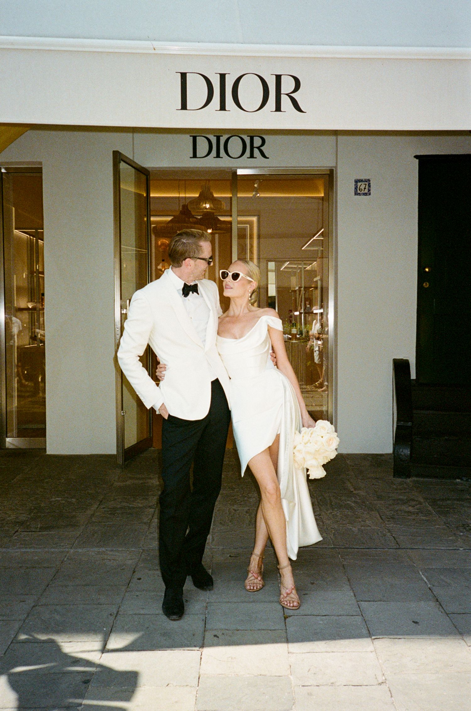 Elegant couple in wedding attire posing outside a Dior boutique, bride in white mini dress, groom in white tuxedo jacket.