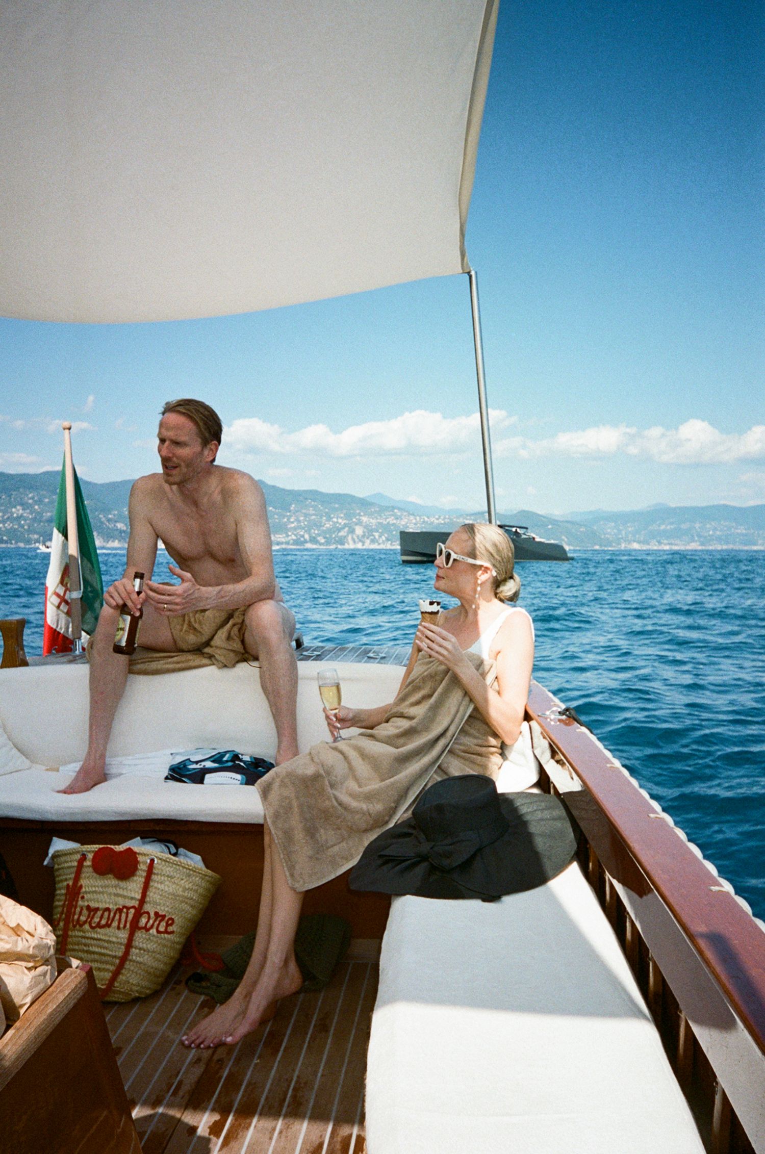Couple relaxing on a luxury boat deck in the Mediterranean, holding drinks with Italian coastline in background.