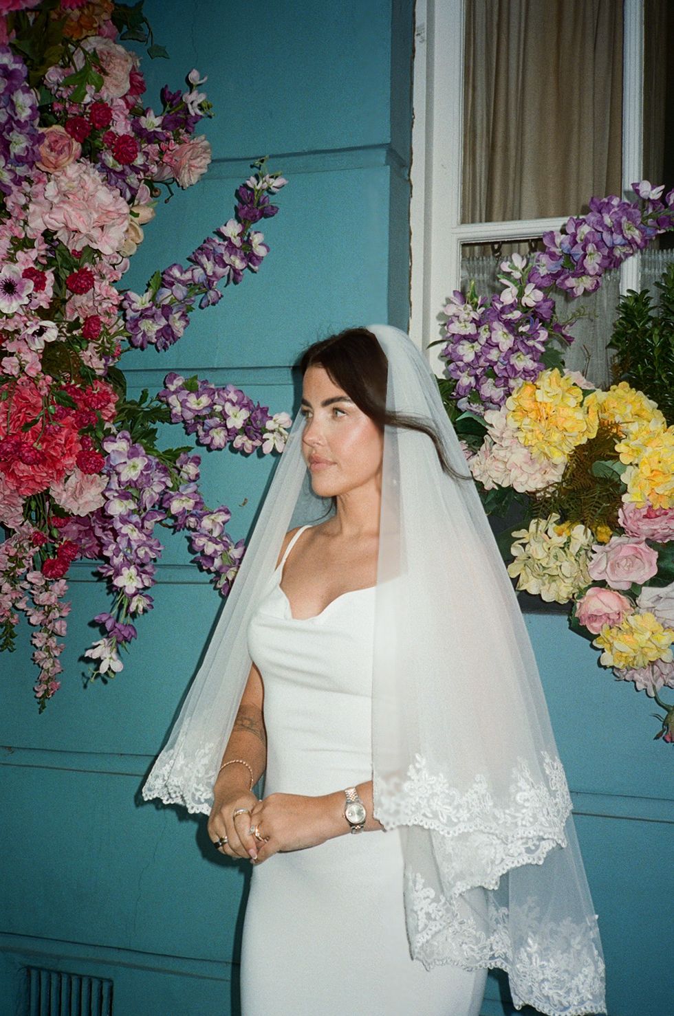 Bride in white gown and lace veil poses before a blue building adorned with colorful floral arrangements.