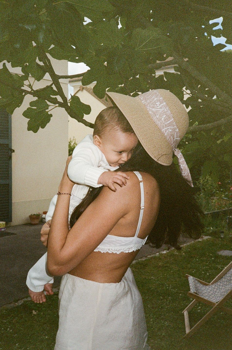 A woman in a white crop top and straw hat lovingly holds a baby outdoors near green trees.