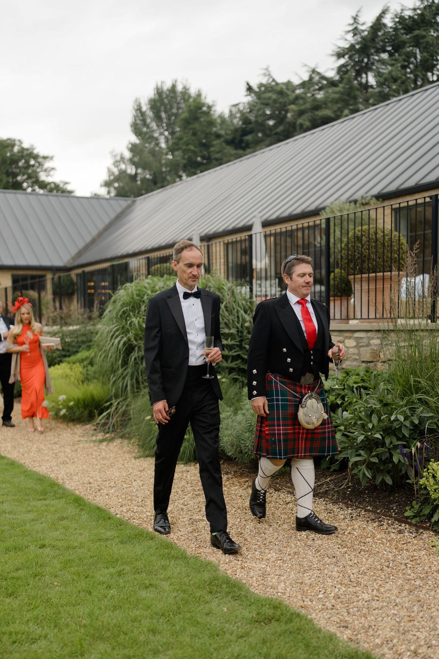 Two men walk at a wedding, one in a black tuxedo and one in traditional Scottish tartan kilt with red tie.