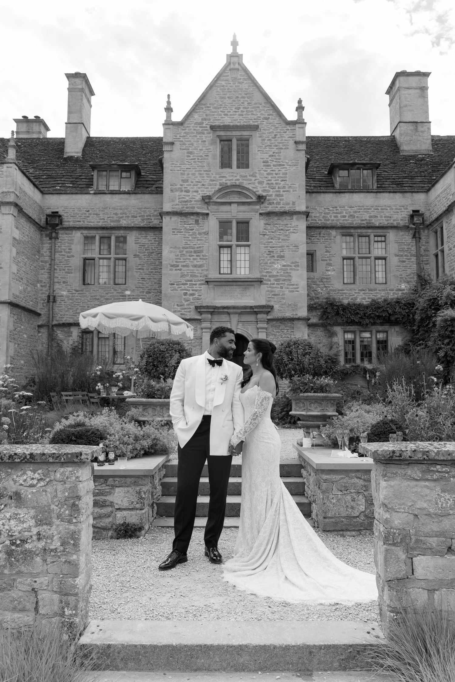 Black and white portrait of bride and groom embracing before grand historic stone manor house.