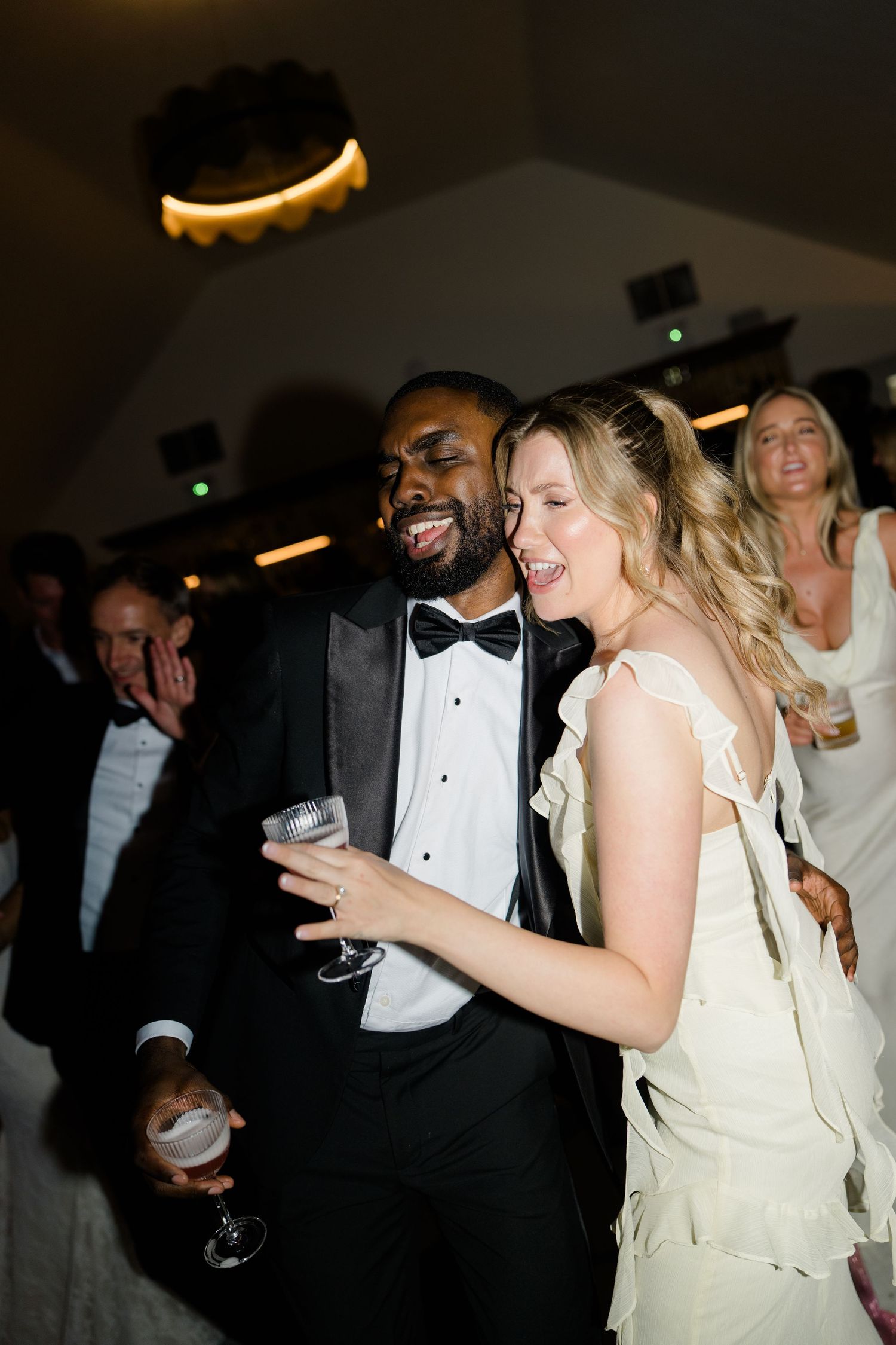 Joyful interracial couple dancing at wedding reception, groom in tuxedo and bride in white dress holding champagne glasses.