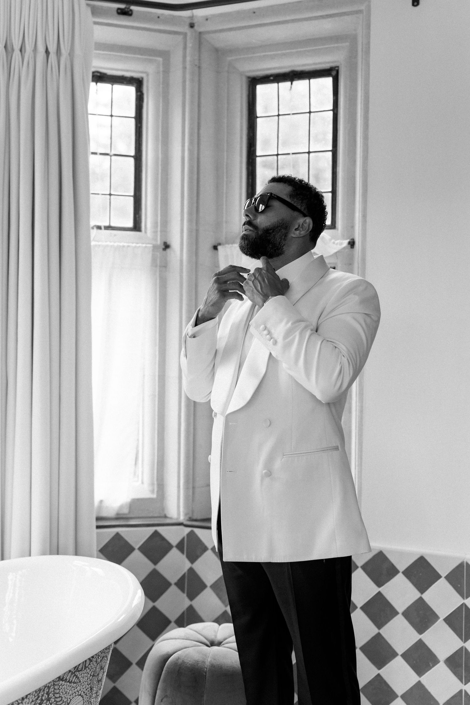 Groom in white tuxedo jacket adjusting bow tie in elegant bathroom with checkered floors.