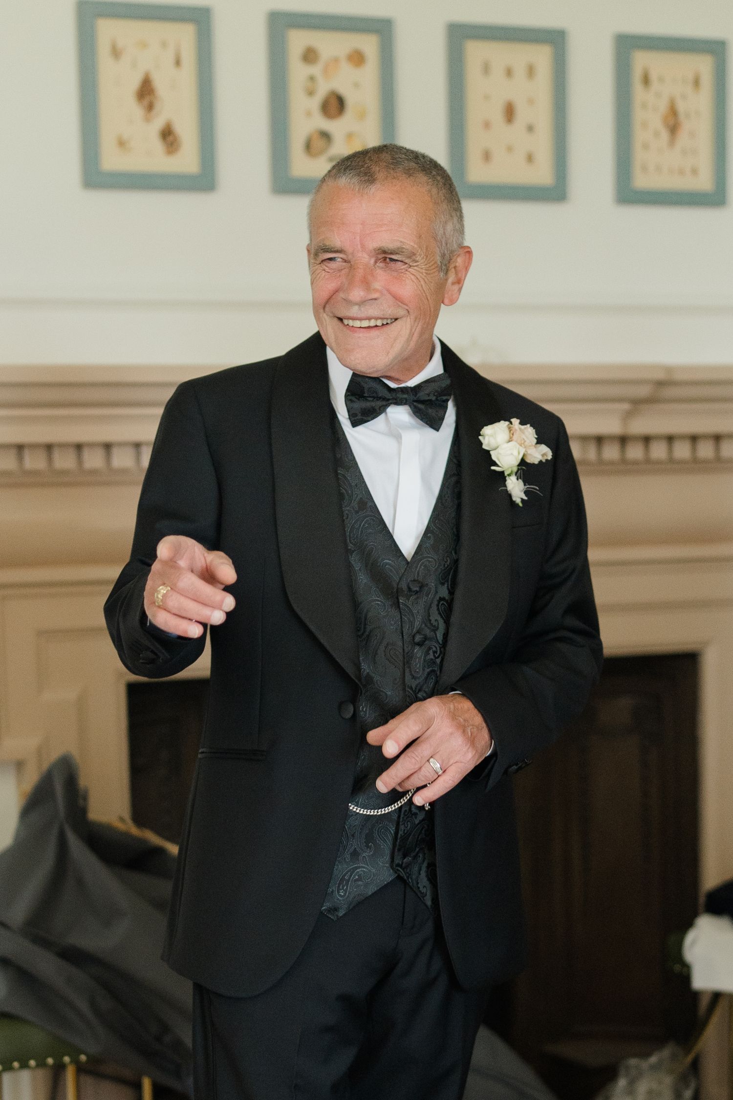 Elegant older man in black tuxedo with boutonniere smiles and points at wedding event.