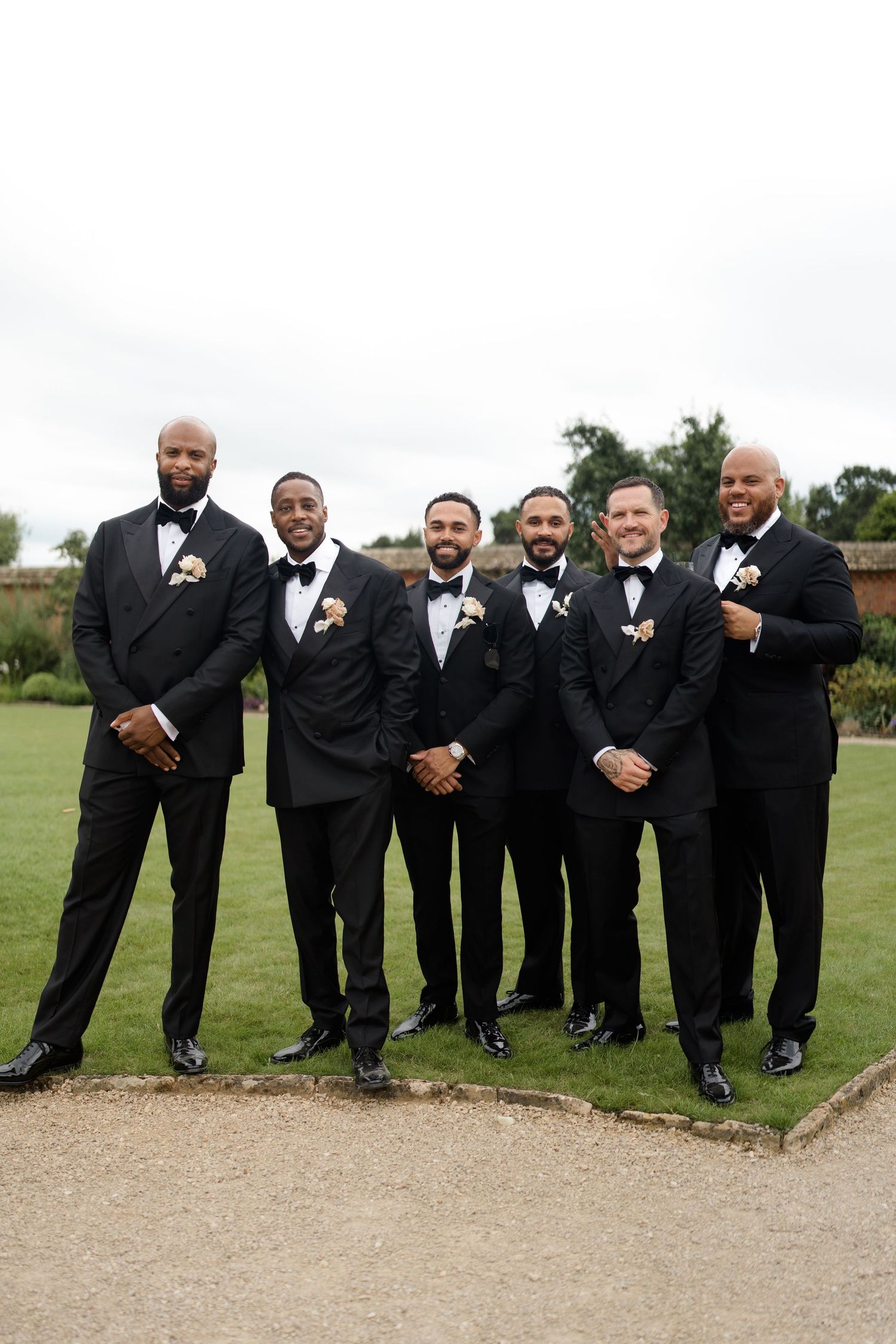Groom and five groomsmen in matching black tuxedos with boutonnieres posing on garden lawn.