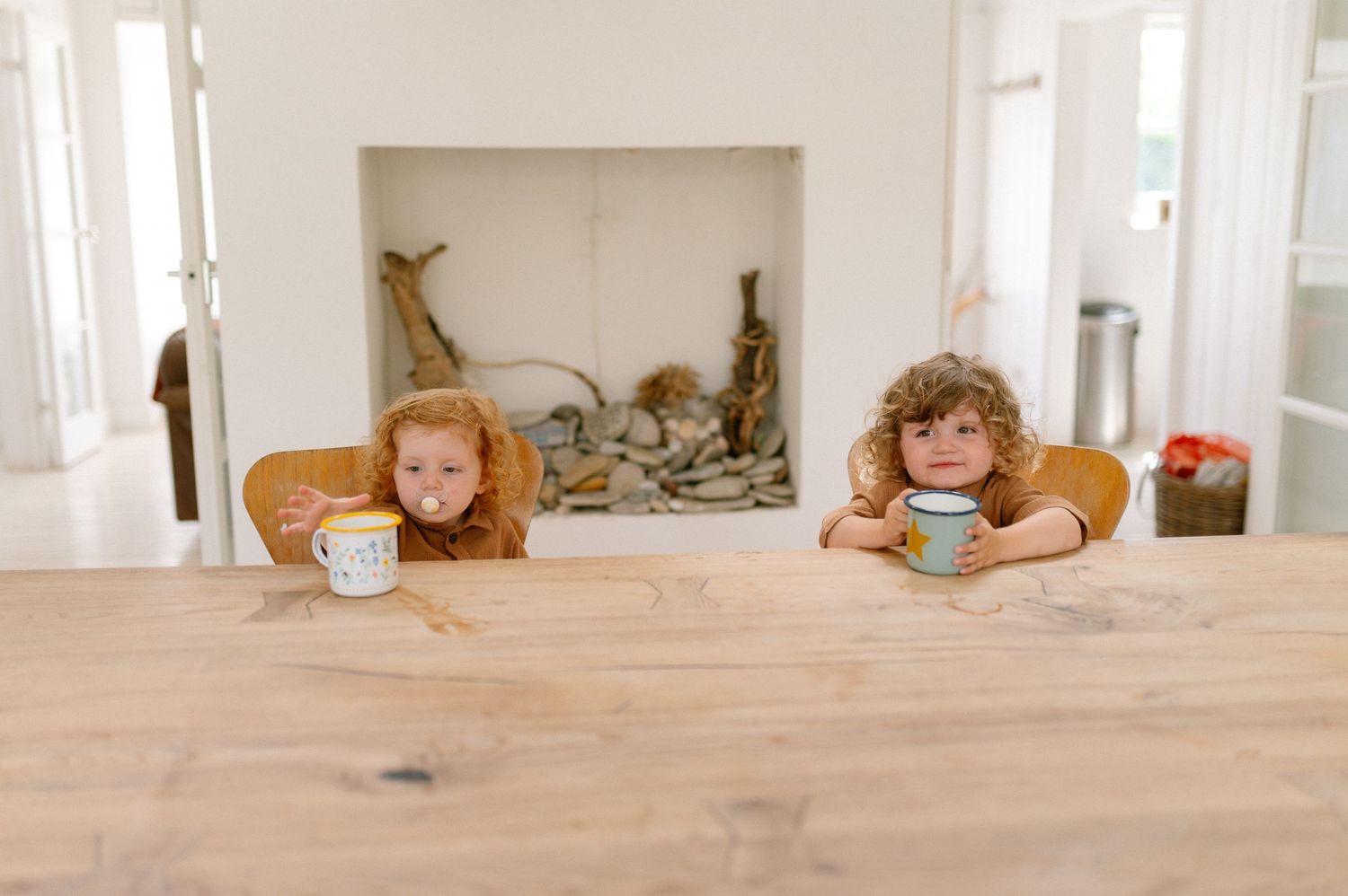 Two coffee mugs sit on a light wooden table in a bright, minimalist interior setting.