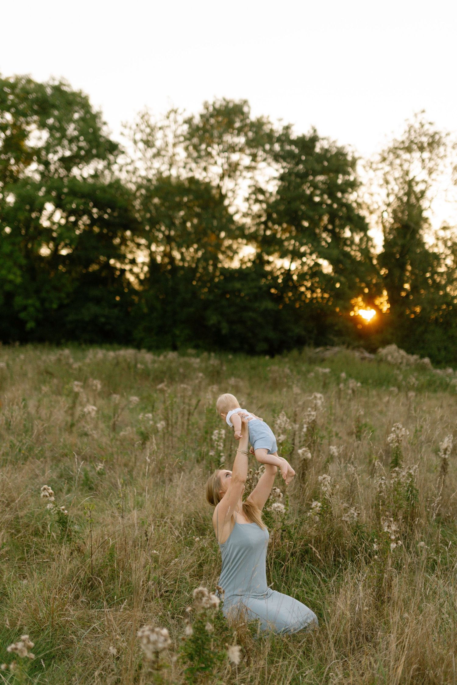 A figure lifts a small child in a grassy meadow during golden hour sunset.