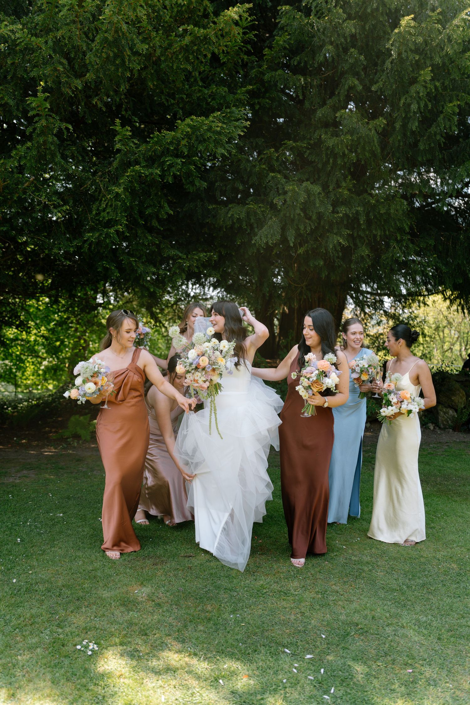 A bridal party in brown and blue dresses poses together outdoors on a sunny day.
