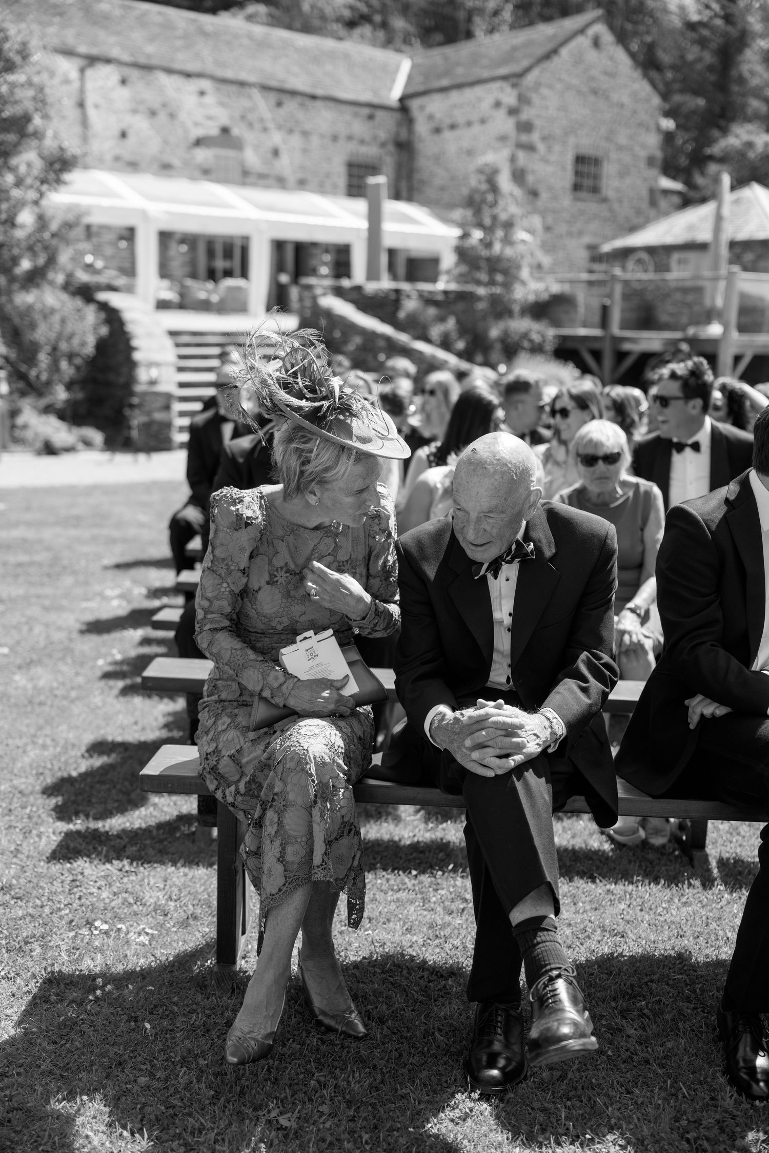 Wedding guests sit on benches outdoors in formal attire on a sunny day.