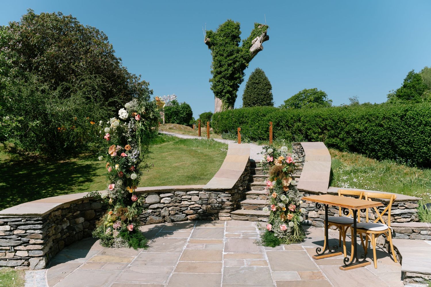 Outdoor wedding ceremony setup with wooden benches and stone walls in a garden setting.