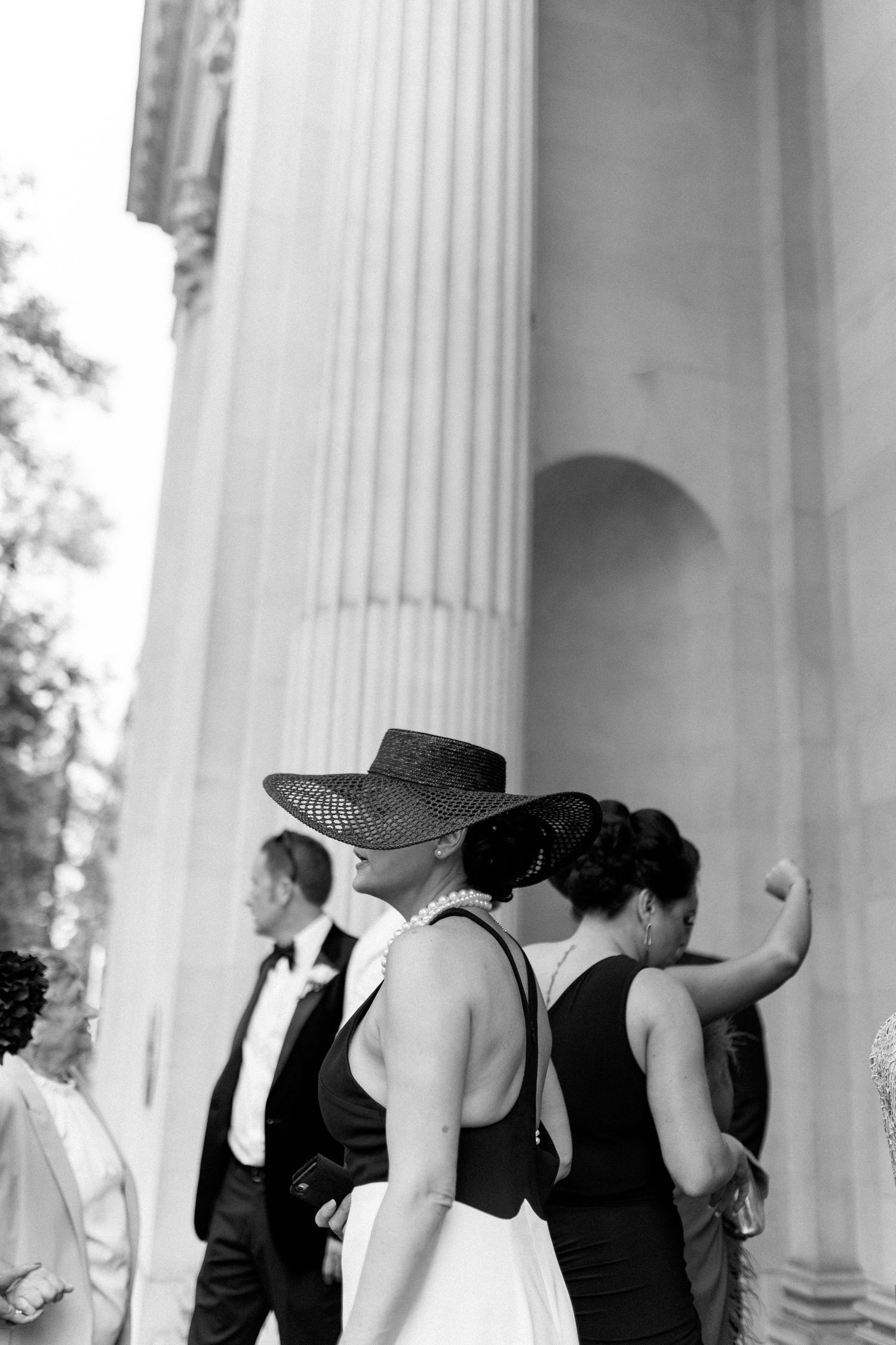 Wedding party in black dresses poses against white stone columns in artistic black and white.