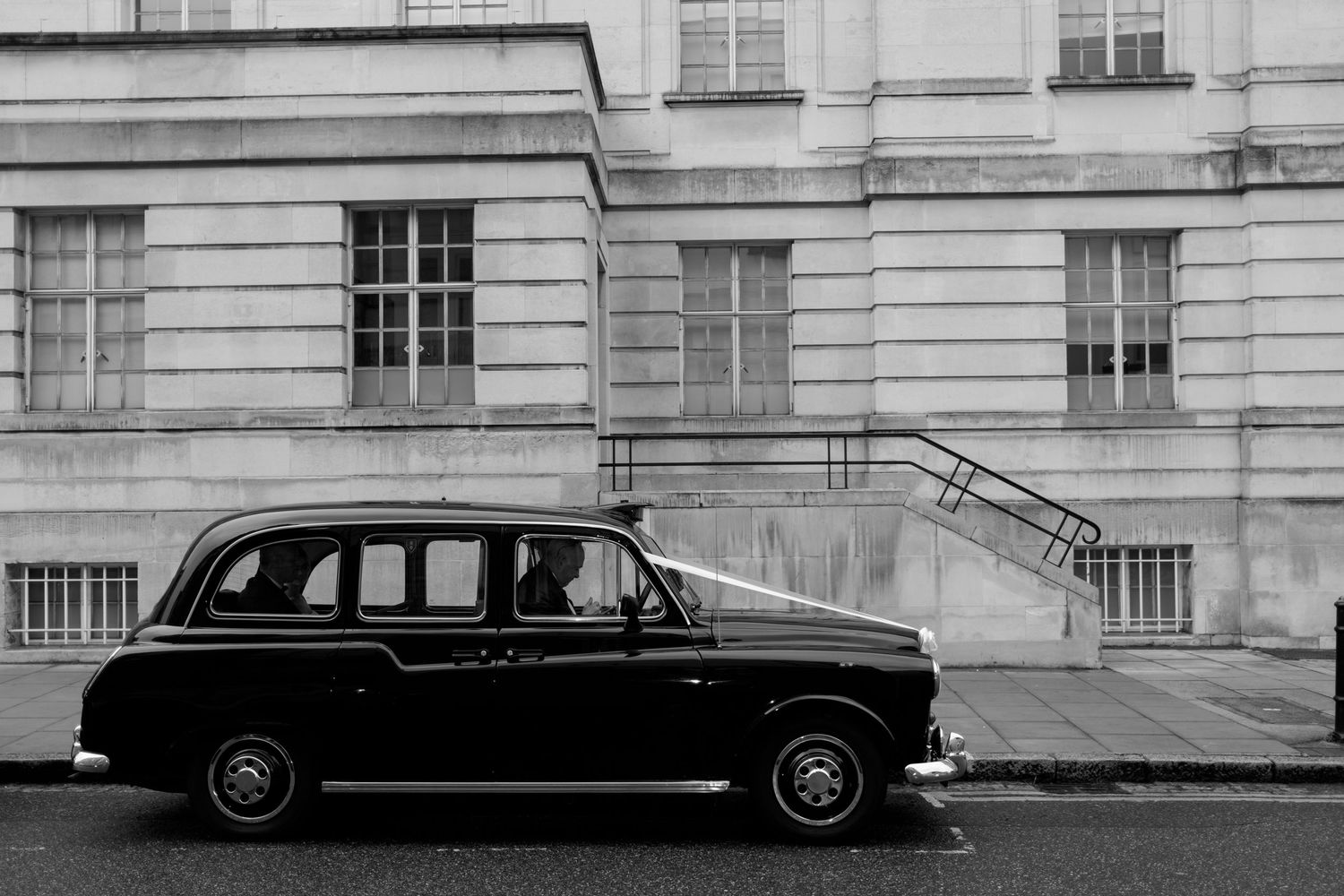 A vintage black taxi cab parked on a street in front of a classic stone building with multiple windows.