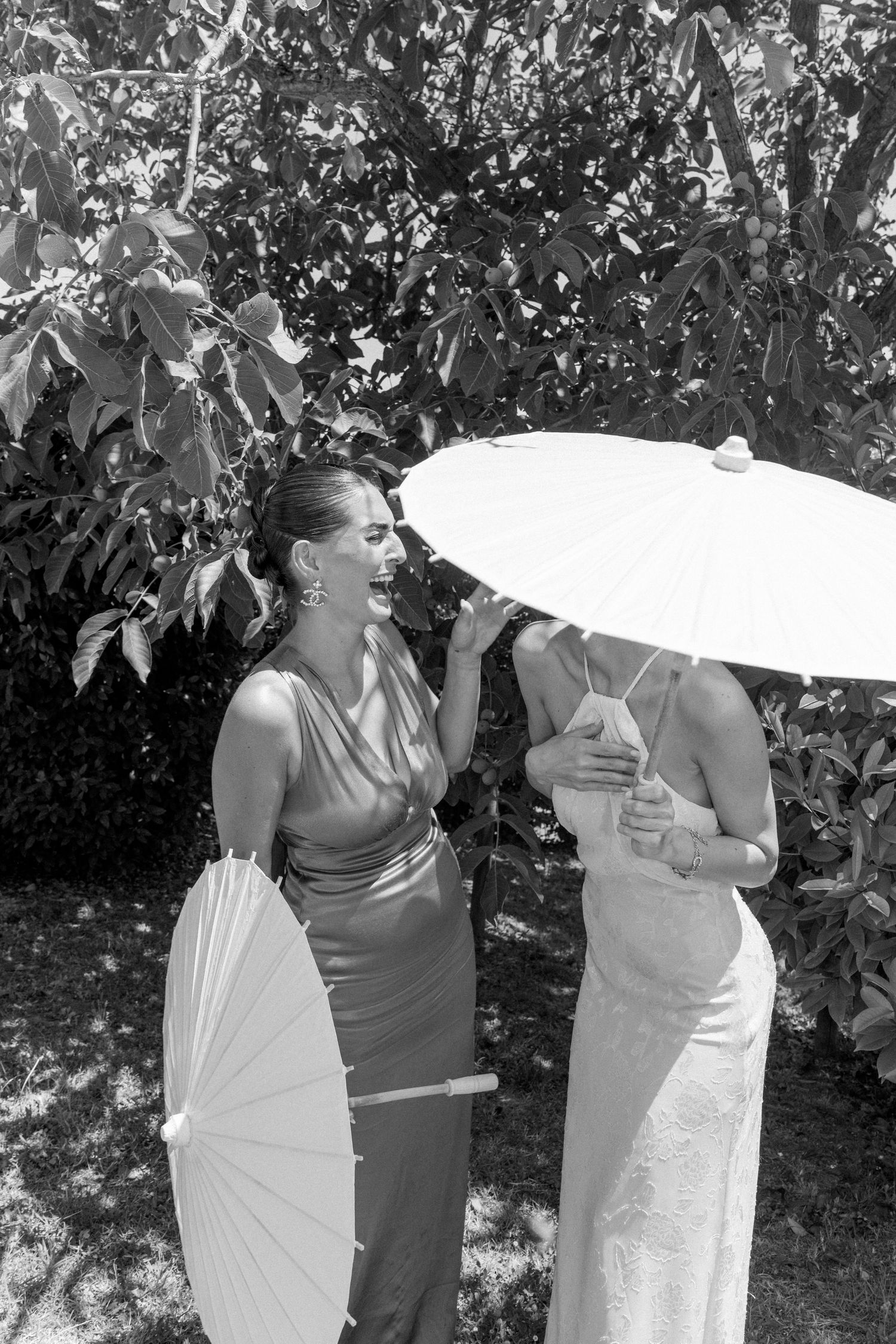 Two people in elegant dresses share a laugh while holding white parasols in a sunny garden setting.