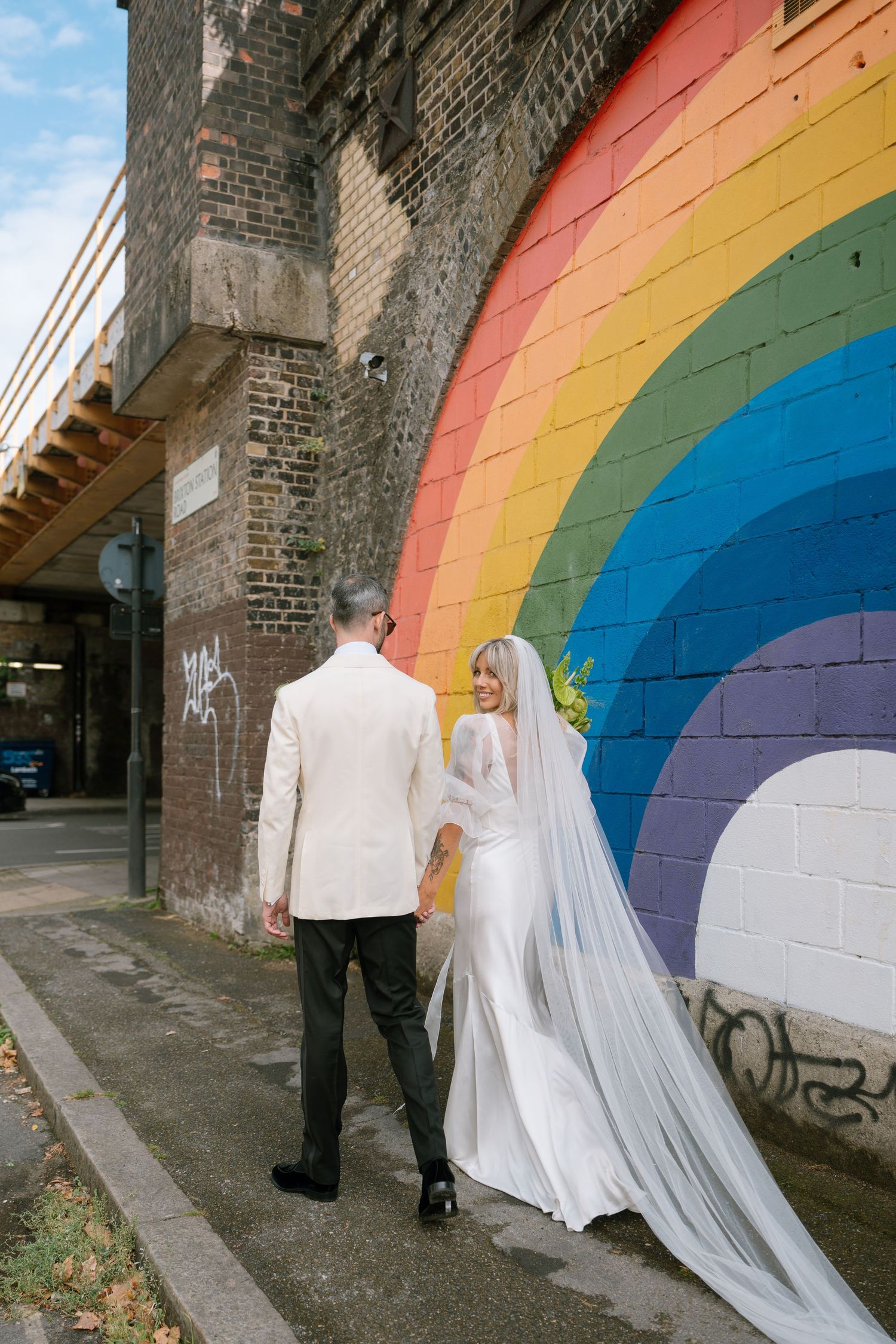 Wedding couple walking along a colorful rainbow mural painted on a brick wall.