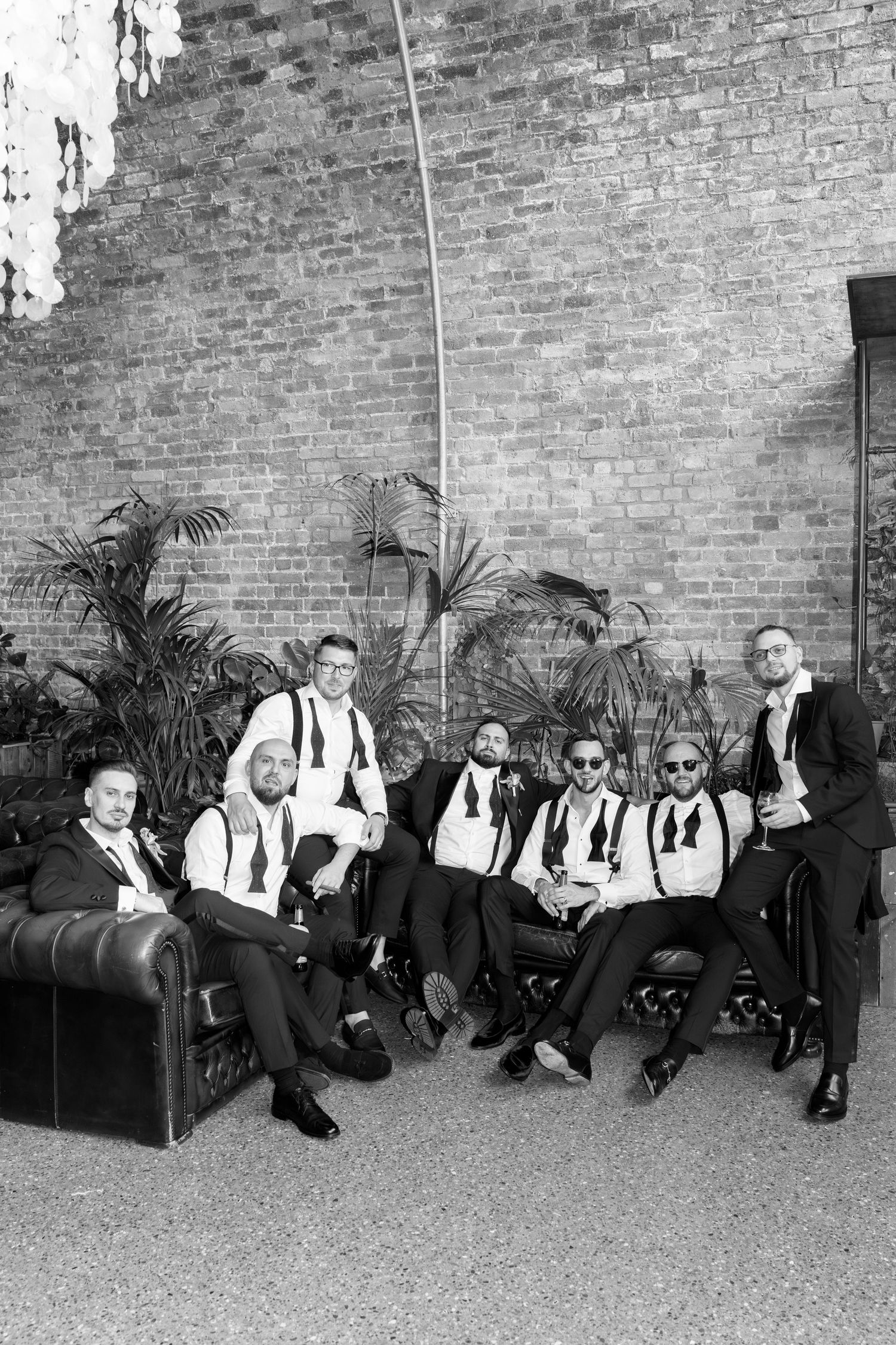 Groomsmen in black suits and suspenders lounging casually against a brick wall.