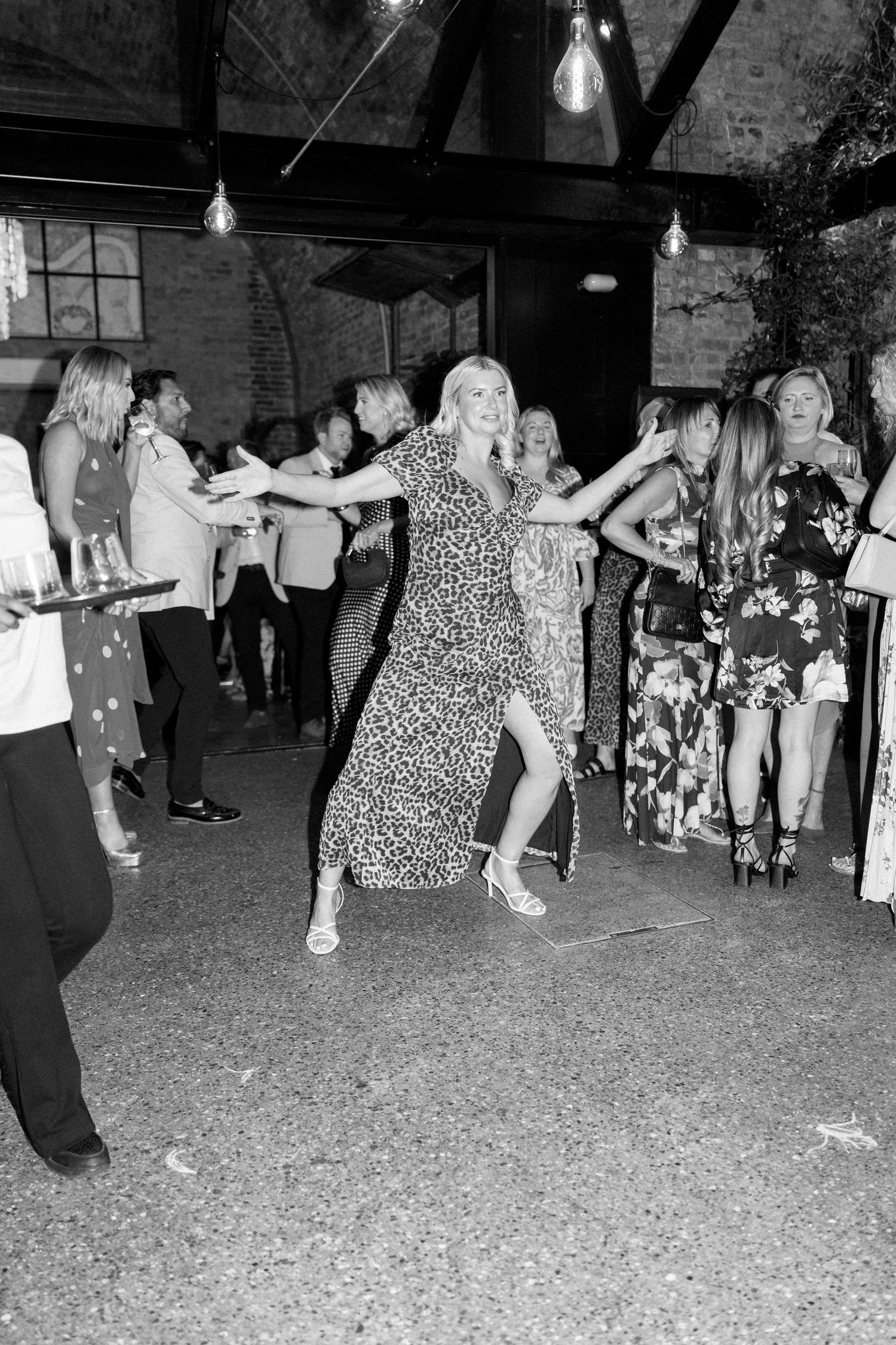 Wedding guests mingle and dance at reception under string lights in industrial venue.