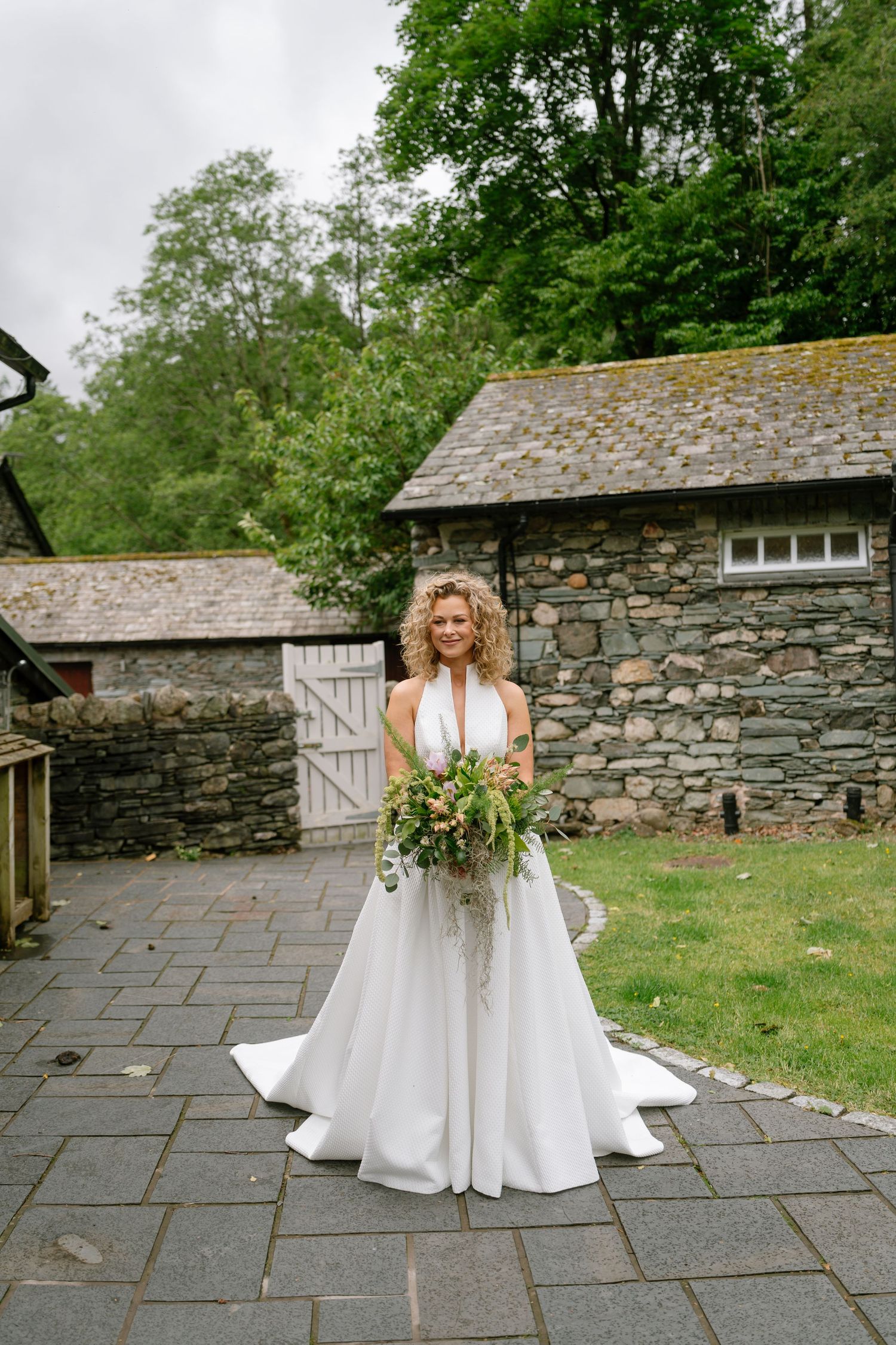 Bride in flowing white dress poses against rustic stone building backdrop.