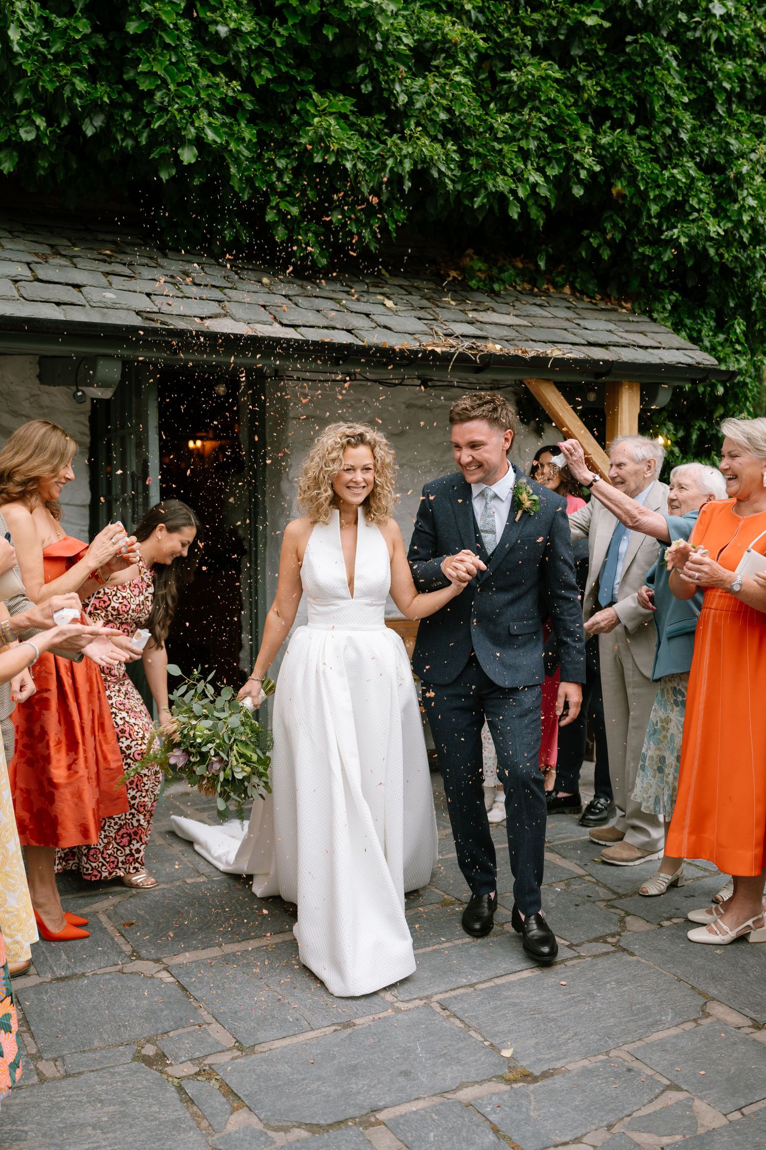Wedding guests in white and orange dresses gather outdoors for a celebratory line dance.