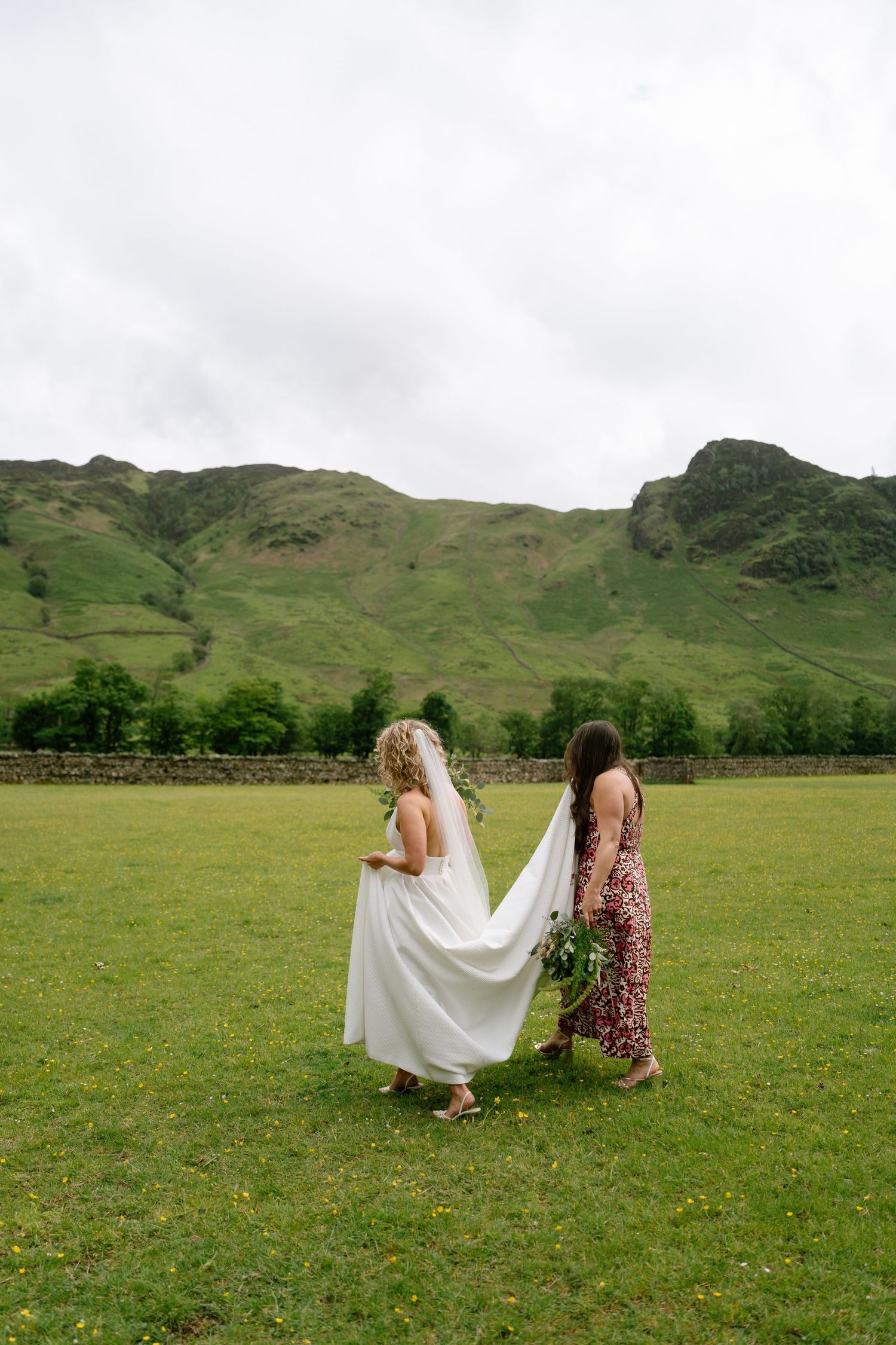 Two people walk across a green field with mountains in the background during a wedding photoshoot.