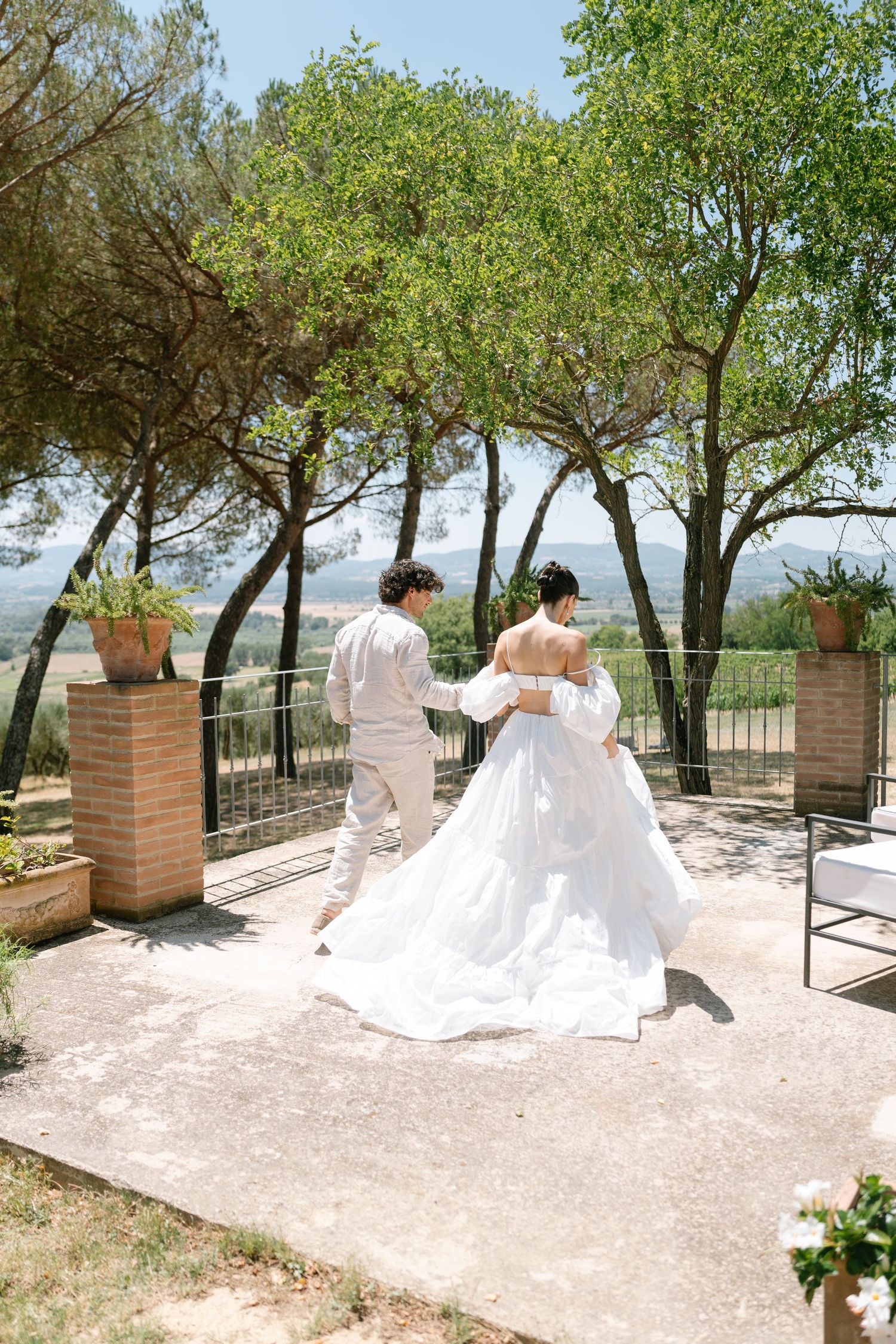 A couple in white attire walking on a terrace surrounded by trees and brick pillars.
