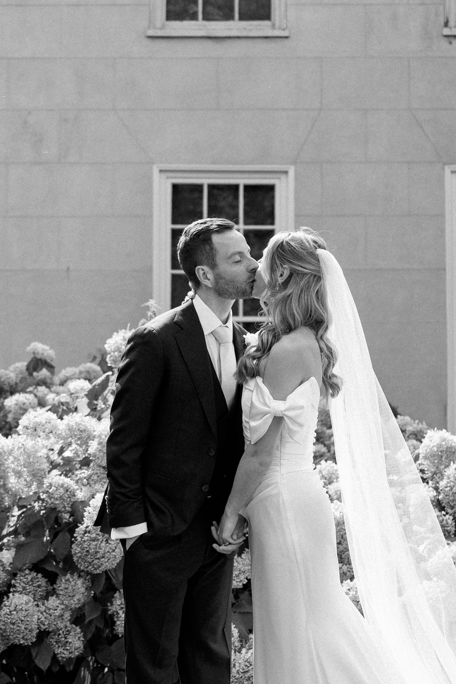 Black and white wedding portrait with hydrangea bushes and classic architecture.