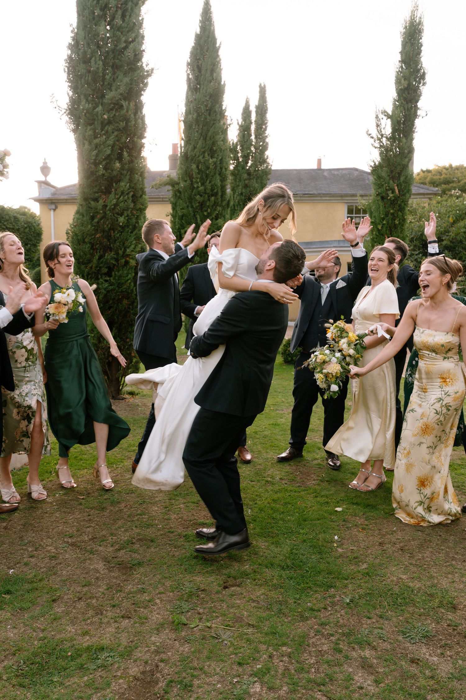 Wedding party performs synchronized celebratory dance moves on grassy lawn near cypress trees.