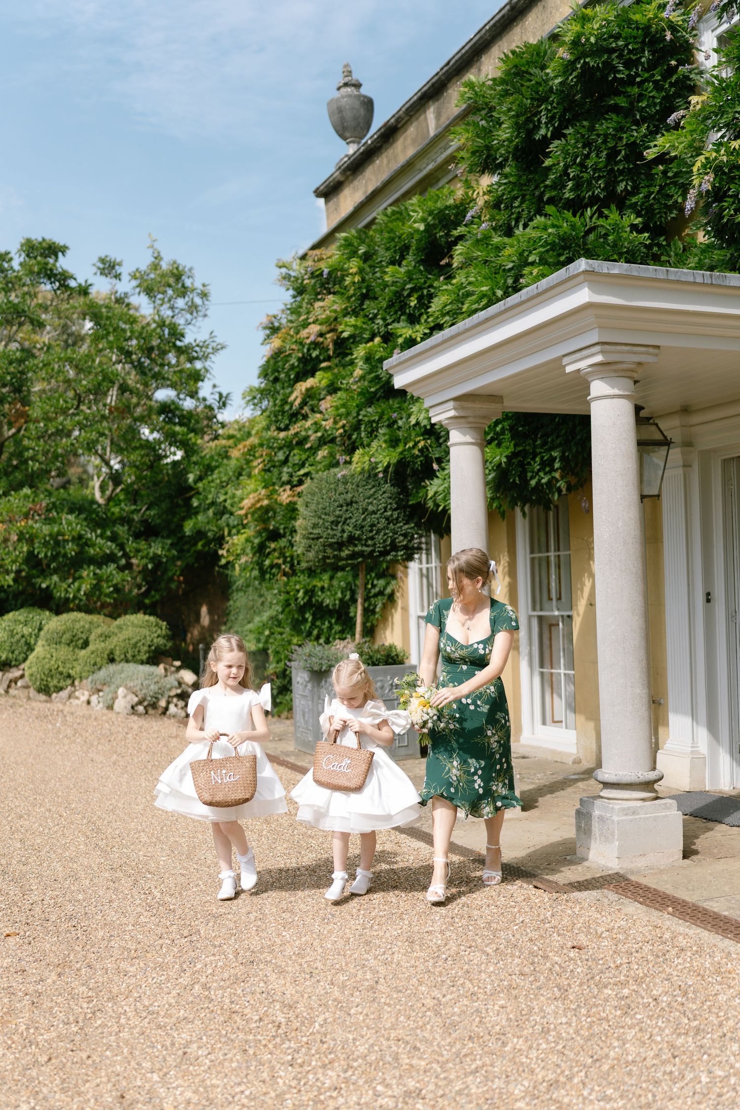 Young attendants in white dresses walking along grand estate entrance with personalized baskets.