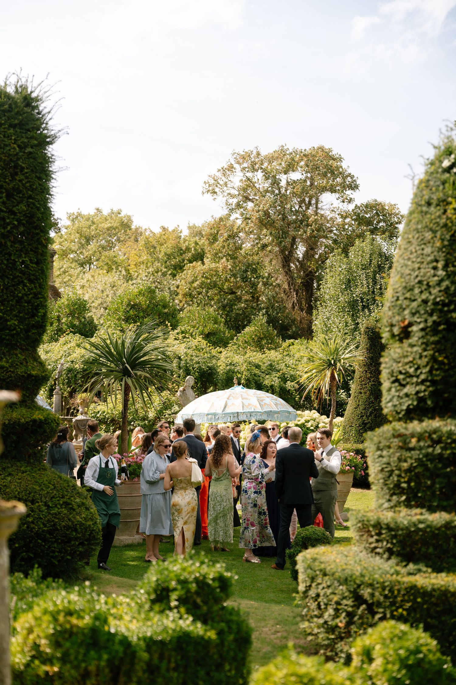 Elegant garden party guests gather under parasols among perfectly manicured topiary and landscaping.