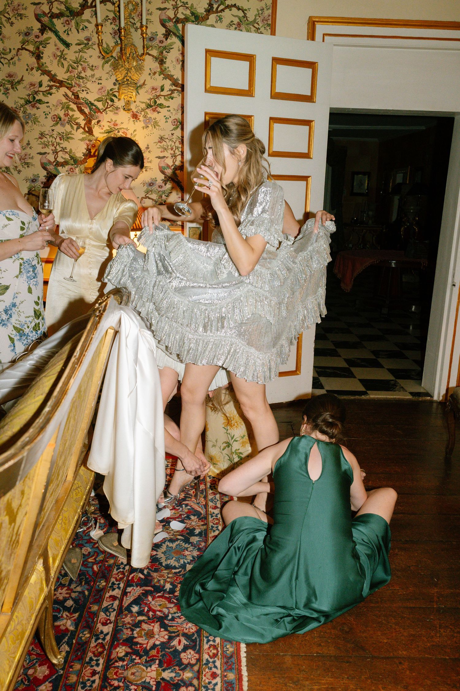 People in silver and green formal attire gathered in an elegant room with floral wallpaper.