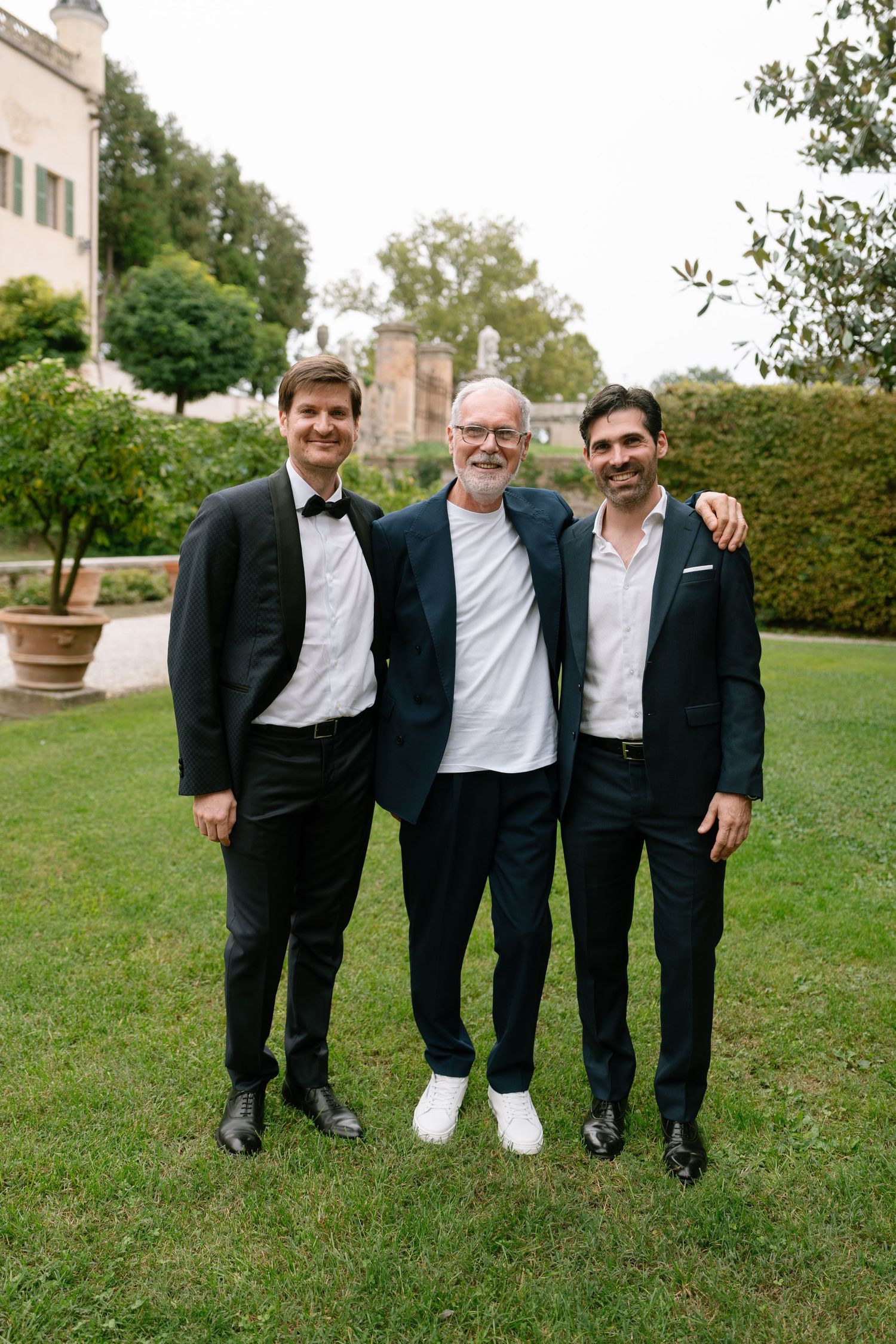 Three people in formal attire pose together on a grassy lawn.