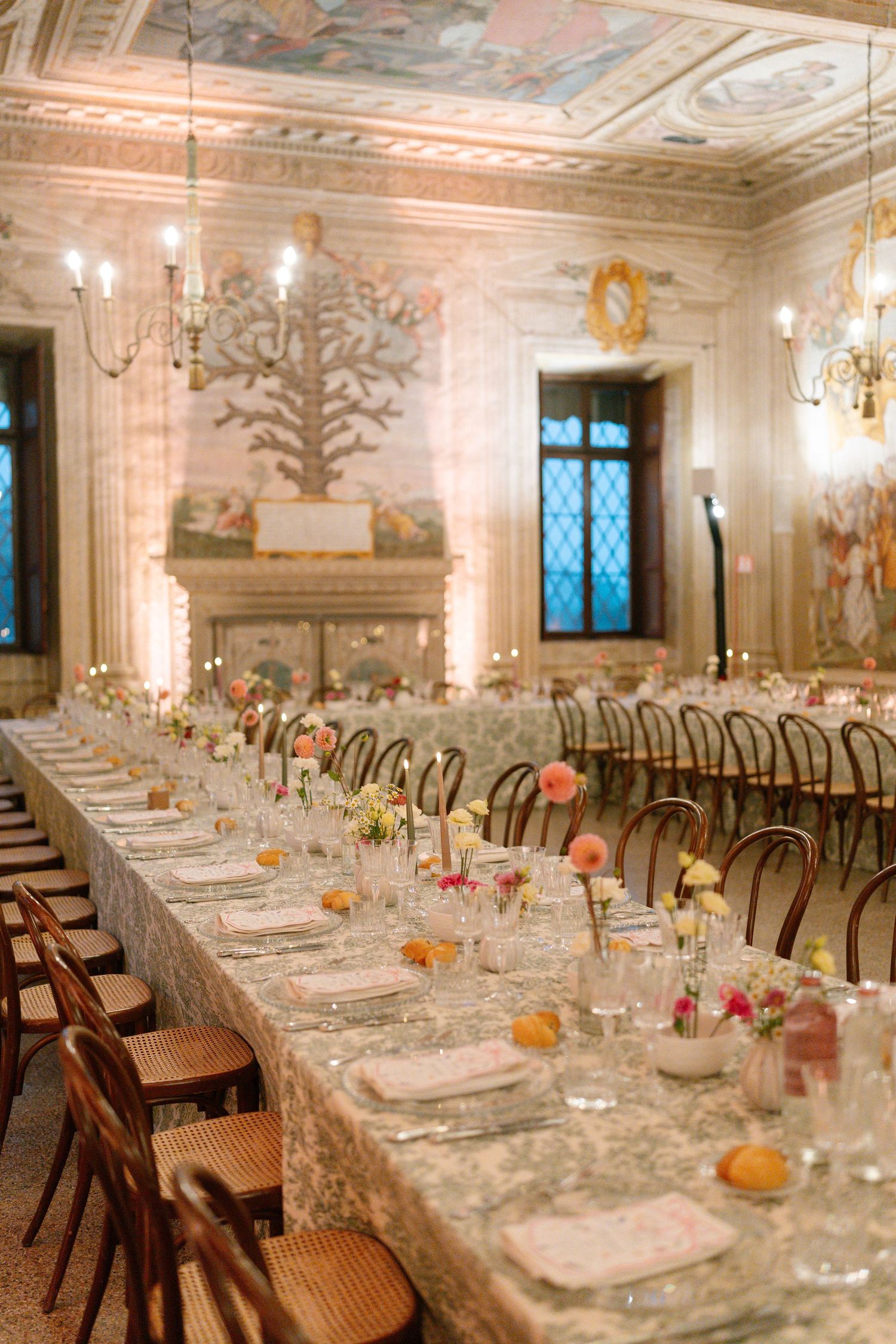 Long banquet tables set with fine china and glassware in an ornate ballroom with painted ceiling murals.