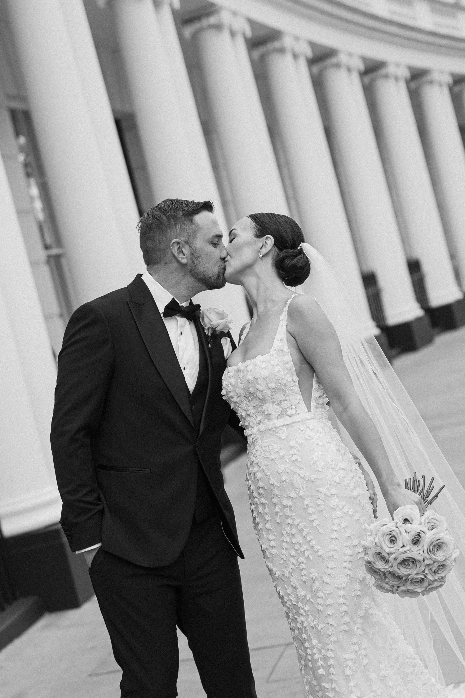 A romantic wedding kiss between newlyweds in front of elegant white columns.