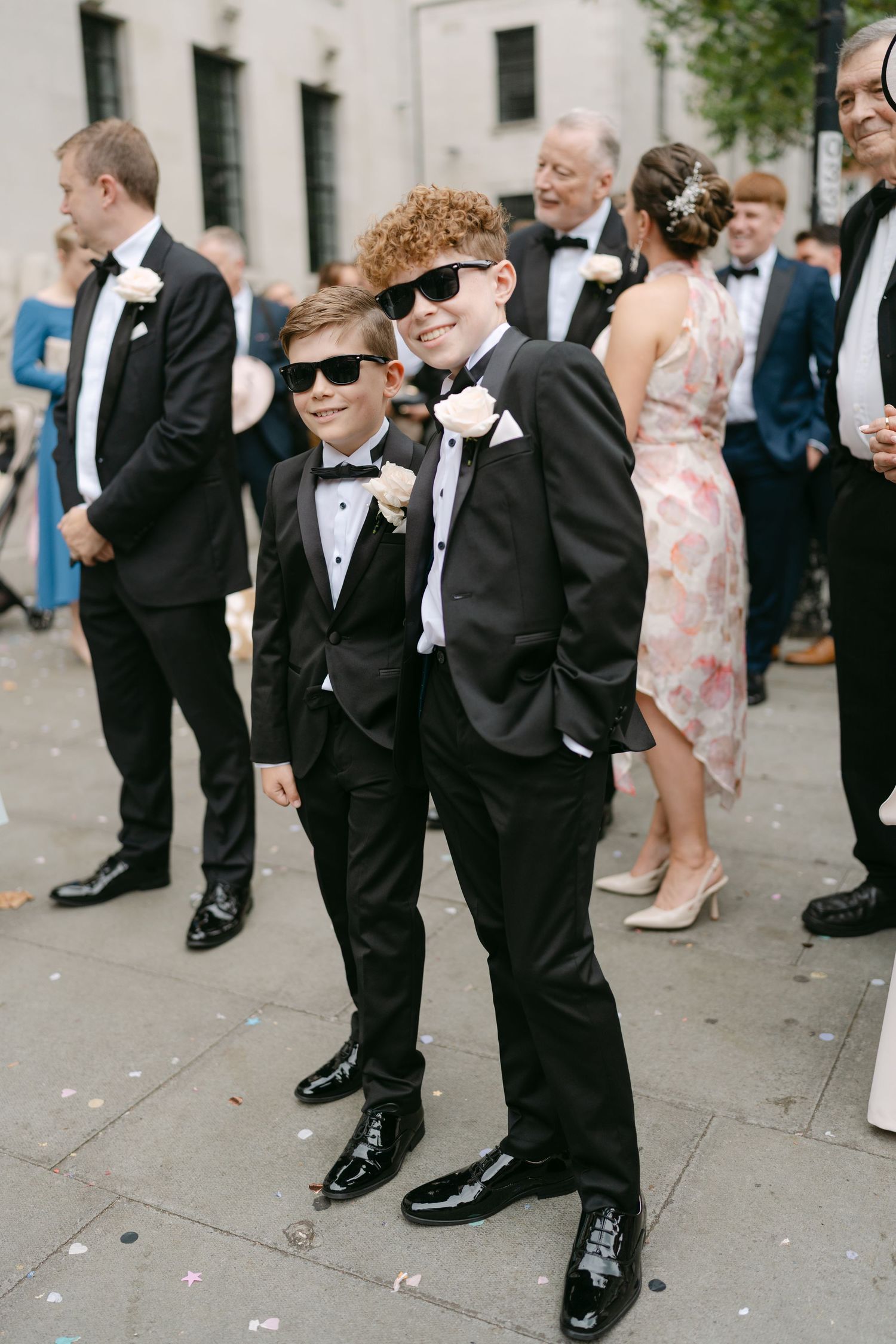 Young groomsmen in tuxedos and sunglasses line up on the sidewalk during a wedding.