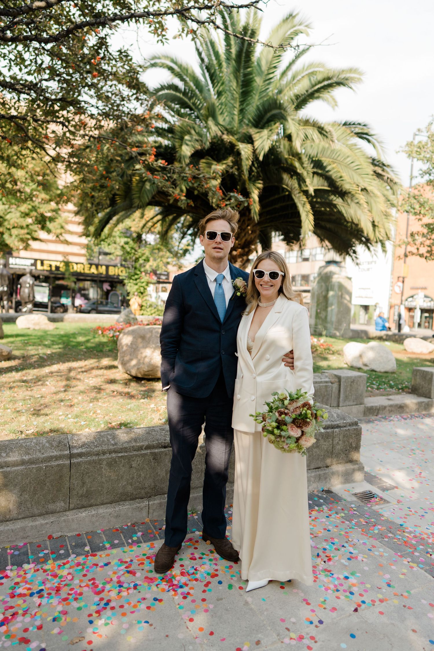 Couple in formal attire pose under palm tree with scattered rose petals at their feet.