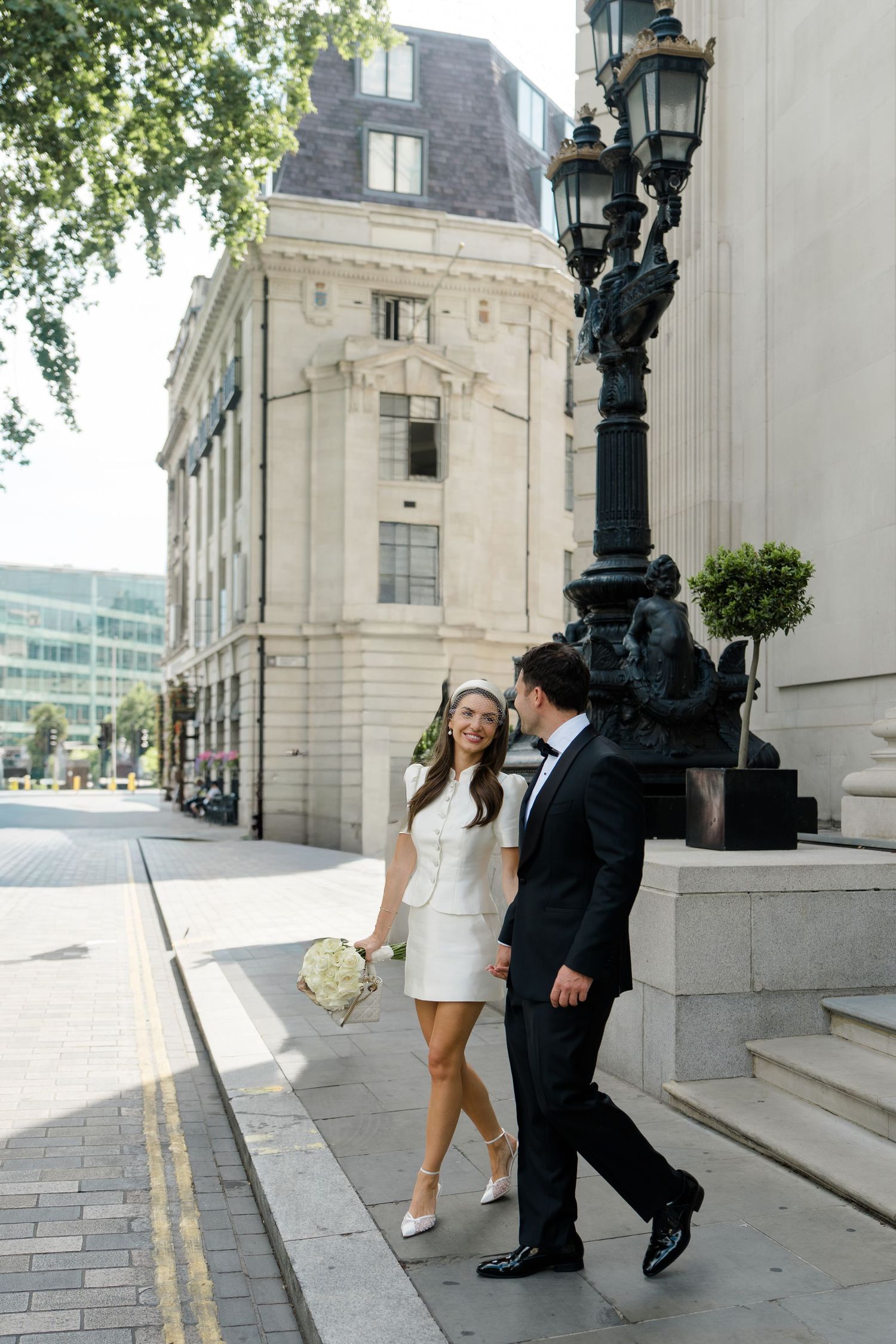 Bride in white mini dress and groom in black tuxedo walking on a London street past a classic lamp post.