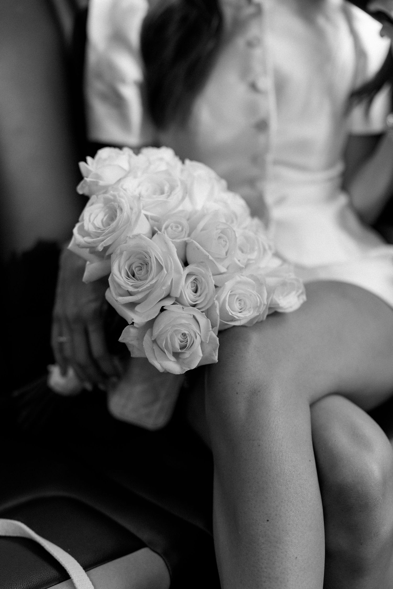 Black and white close-up of bride holding a round white rose bouquet while seated in a car.