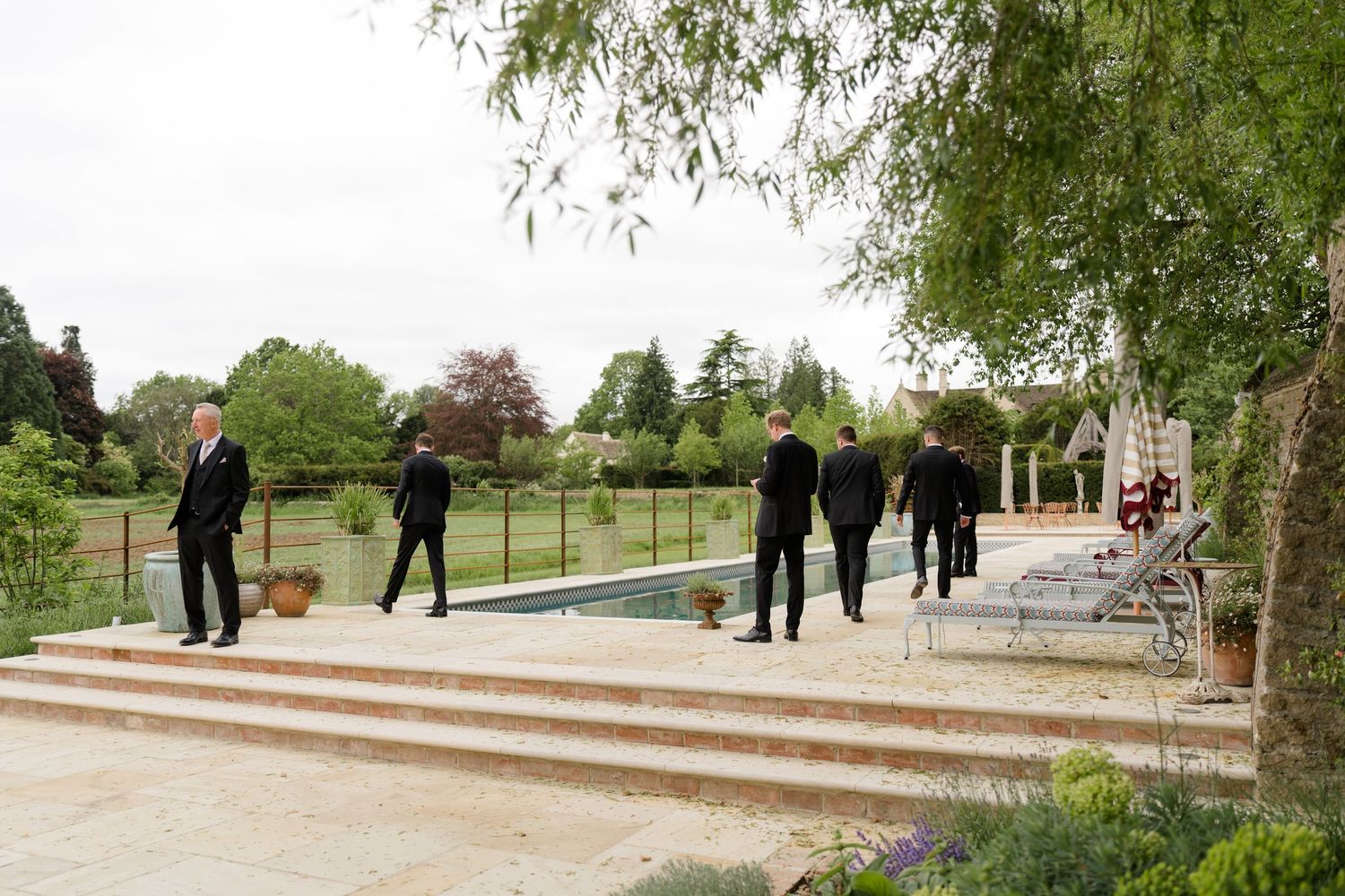 Groomsmen in black tuxedos walk along a stone terrace beside a pool at an elegant garden wedding venue.
