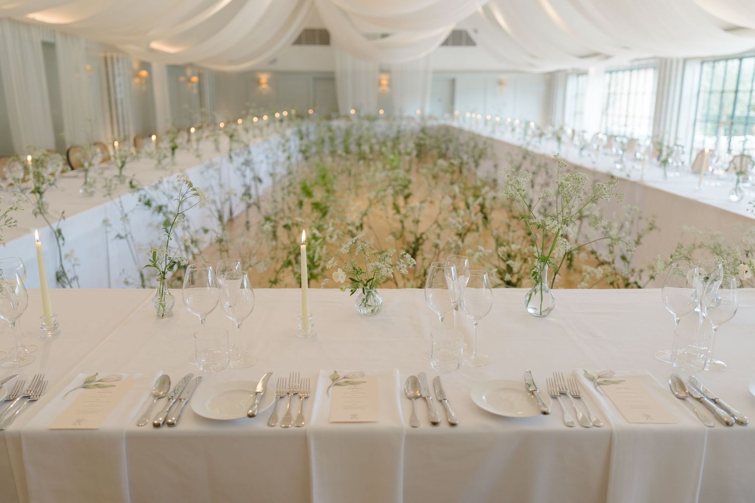 Elegant wedding reception hall features long white tables with candles and floral centerpieces.