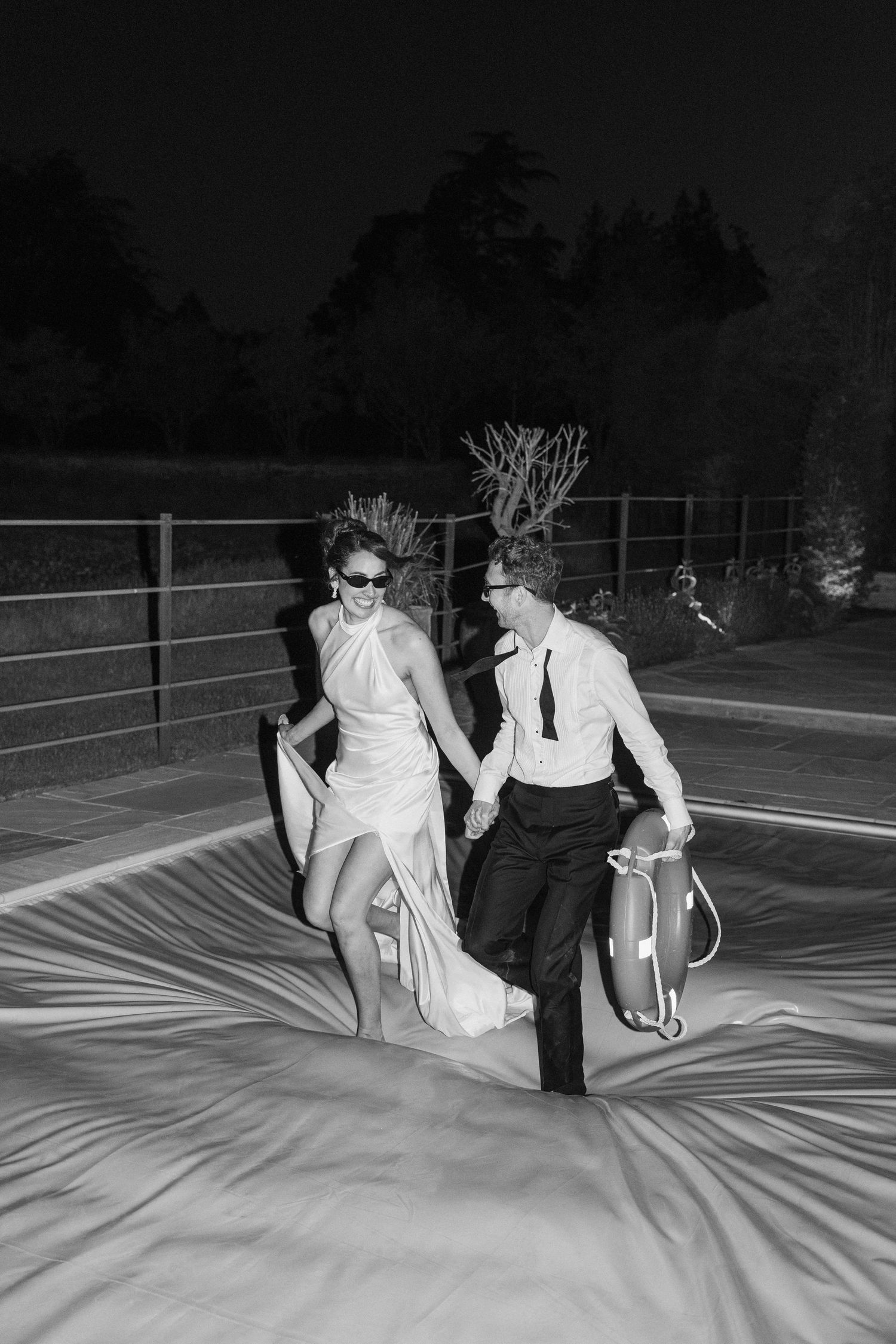 Bride and groom hold life rings laughing on bouncy castle at night reception.