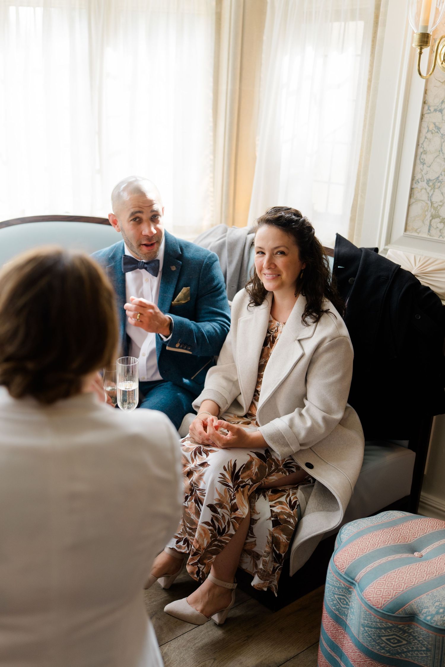 Couple in wedding attire sits on sofa conversing with guest in bright, elegant room.