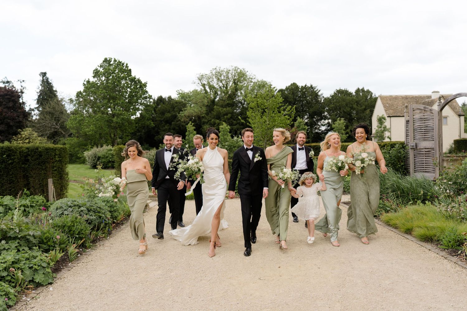 Wedding party walks garden path, bridesmaids in sage gowns, groomsmen in tuxedos at English countryside venue.