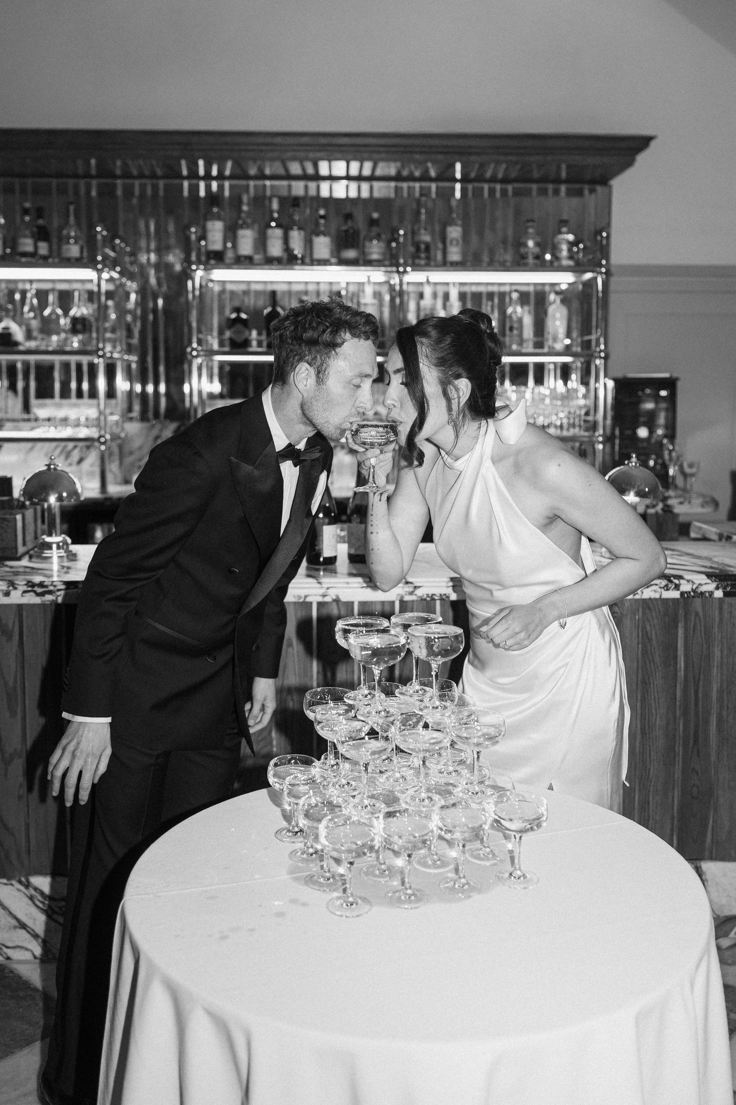Bride and groom sip champagne together beside a champagne tower at their wedding reception, black and white.