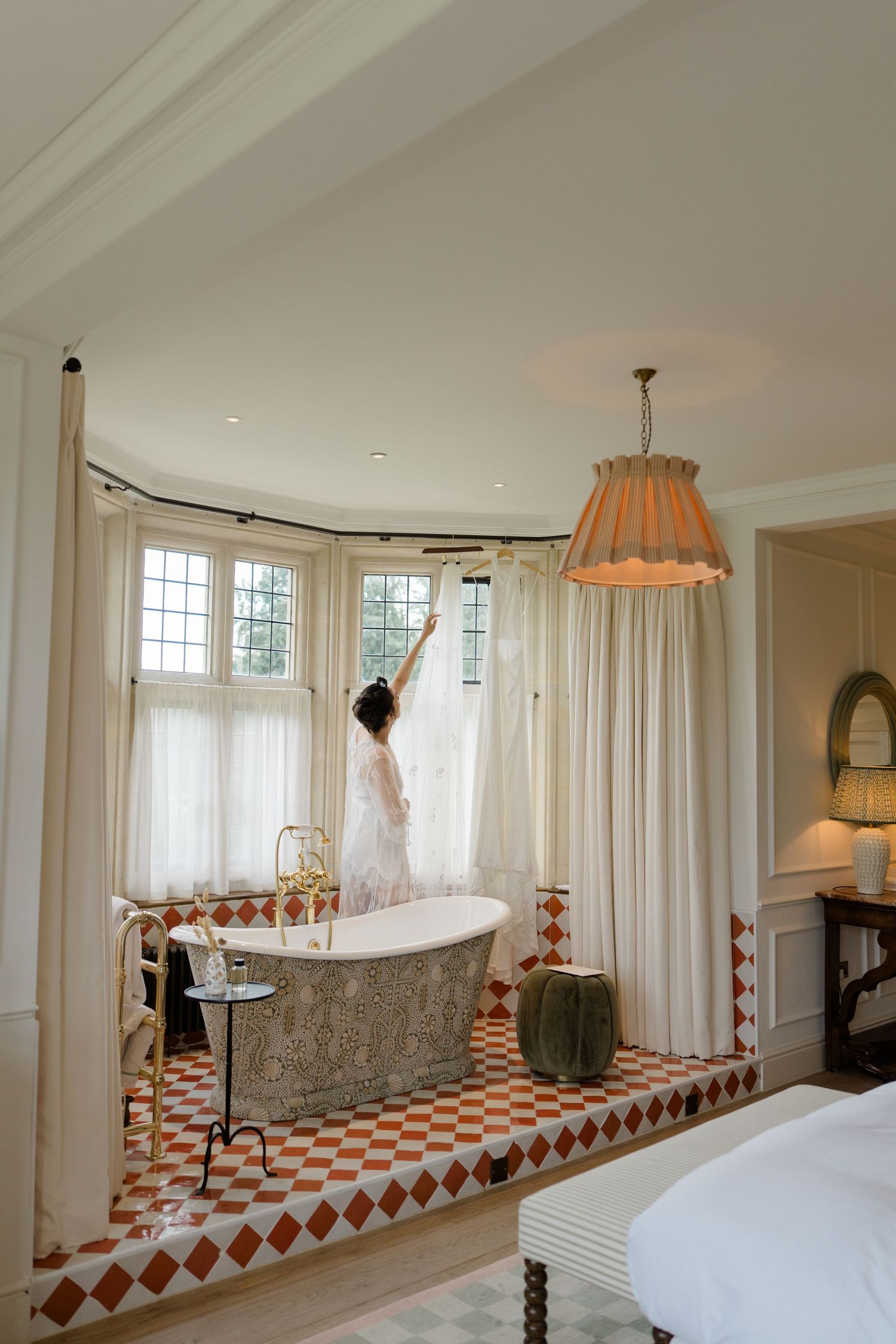 Bride in white robe reaching up to open curtains beside a freestanding ornate bathtub on diamond tile platform.