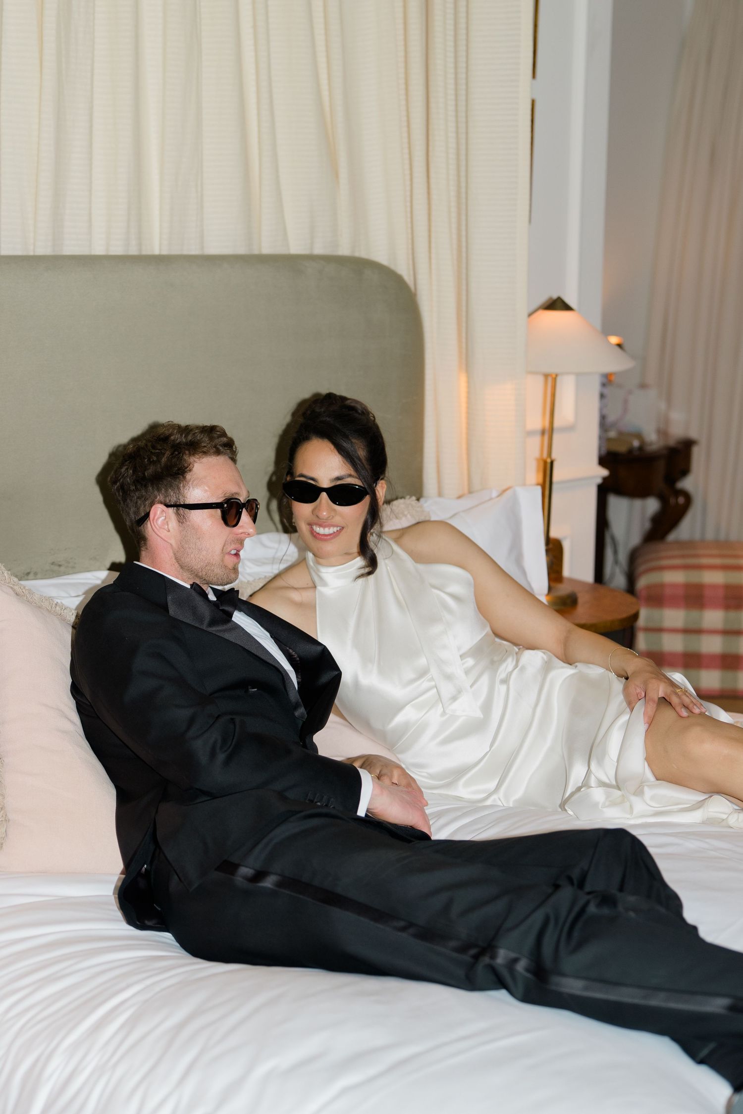 Bride and groom in sunglasses relaxing together on hotel bed in wedding attire after ceremony.