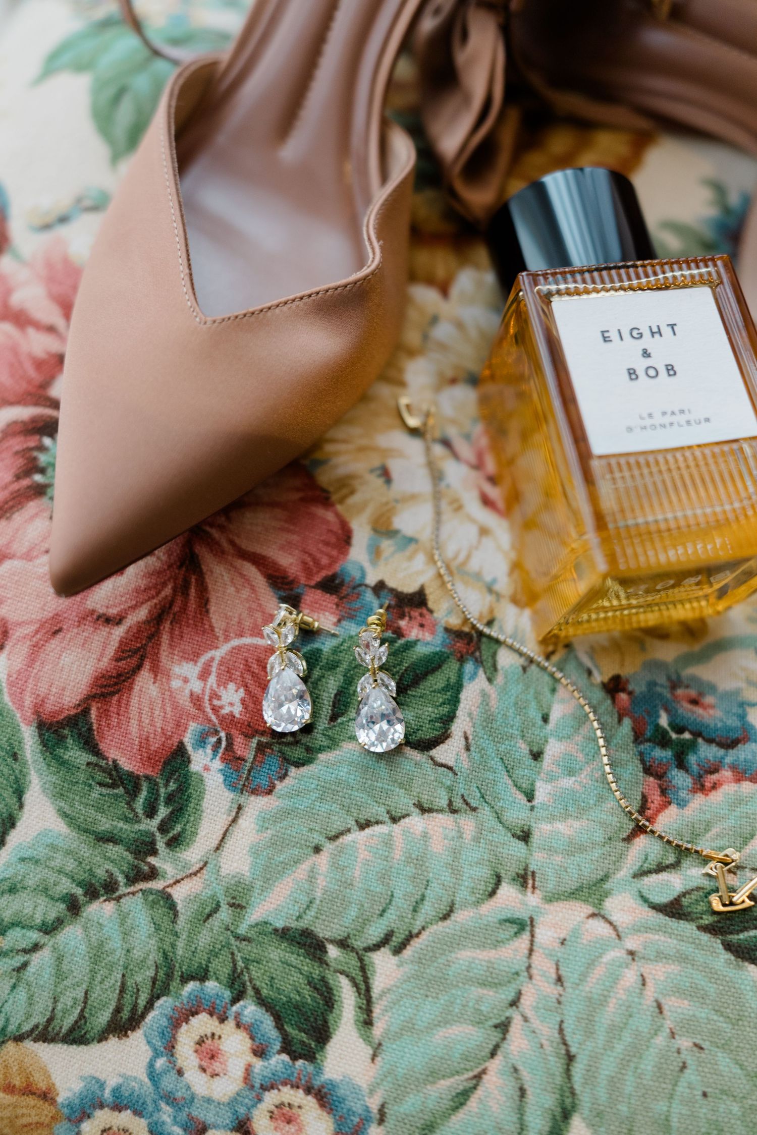 Bridal flat lay with nude heels, crystal drop earrings, gold bracelet, and Eight and Bob perfume on floral fabric.
